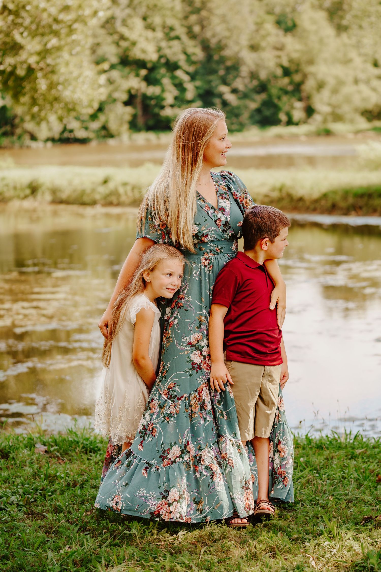 Blonde mother in floral dress with two children by peaceful pond, summer family portrait.