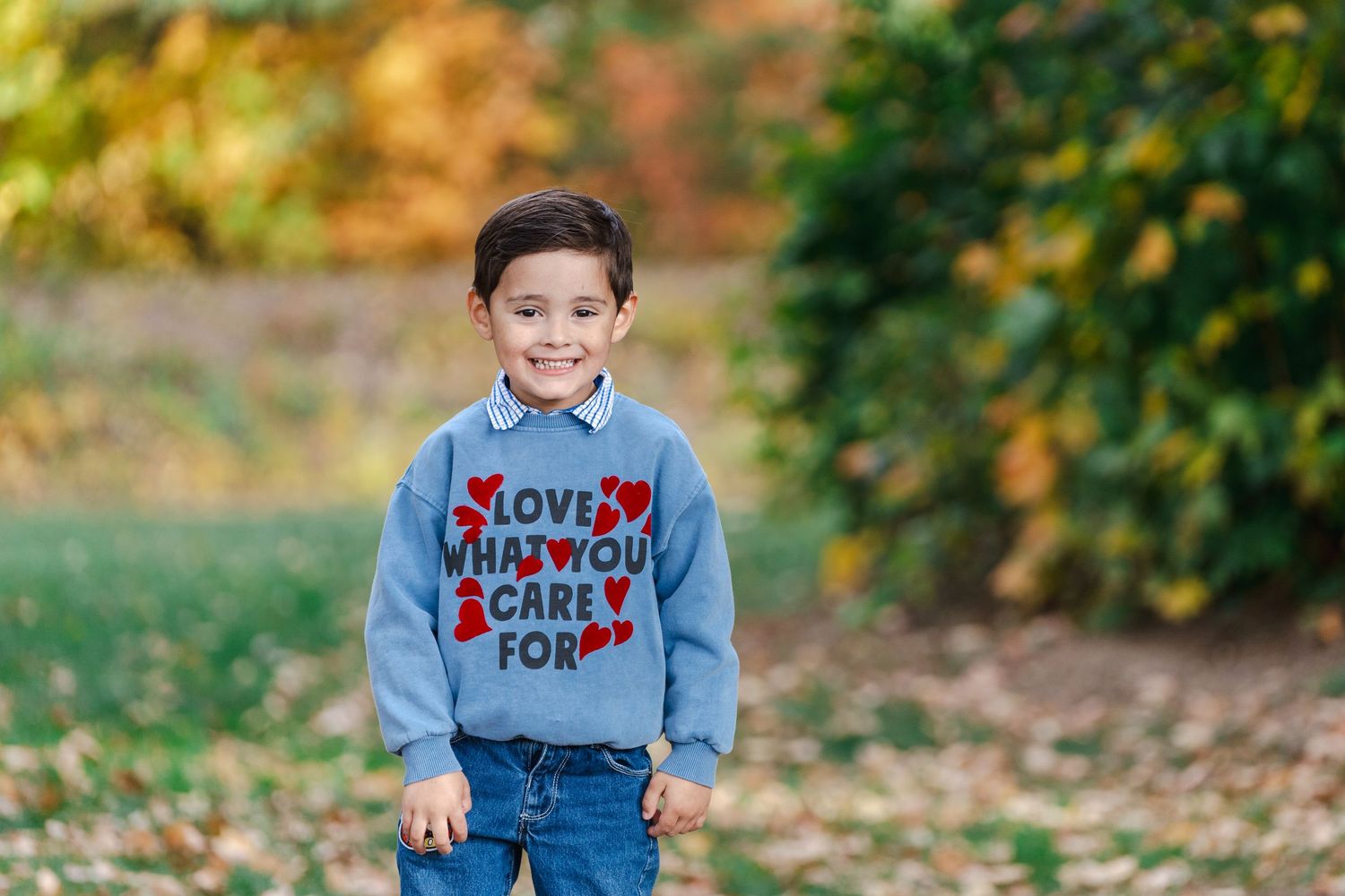Smiling young boy wearing a blue love sweater and jeans standing outdoors in a colorful autumn park.