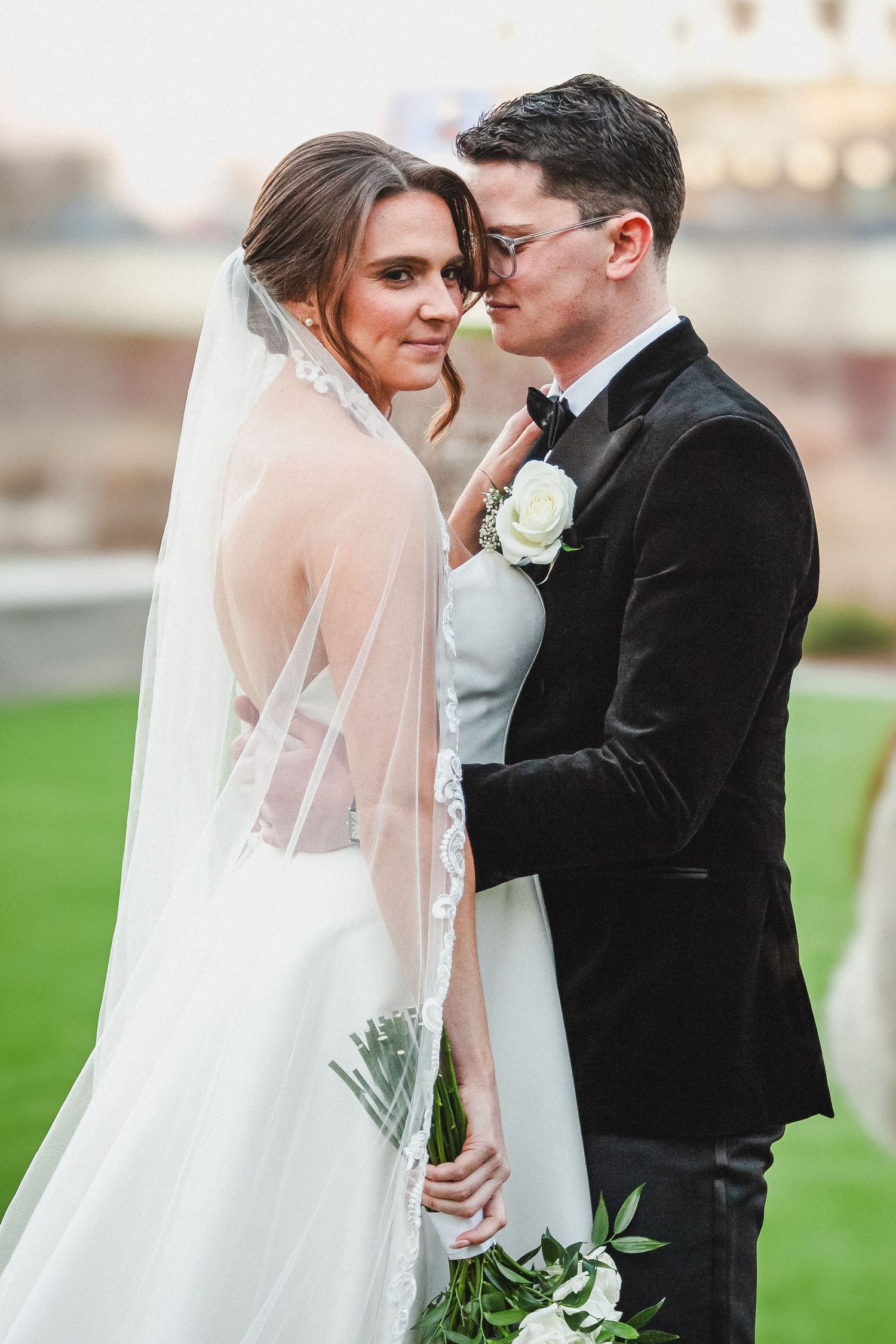 Elegant bride in white gown and veil poses with groom in black tuxedo outdoors at dusk.