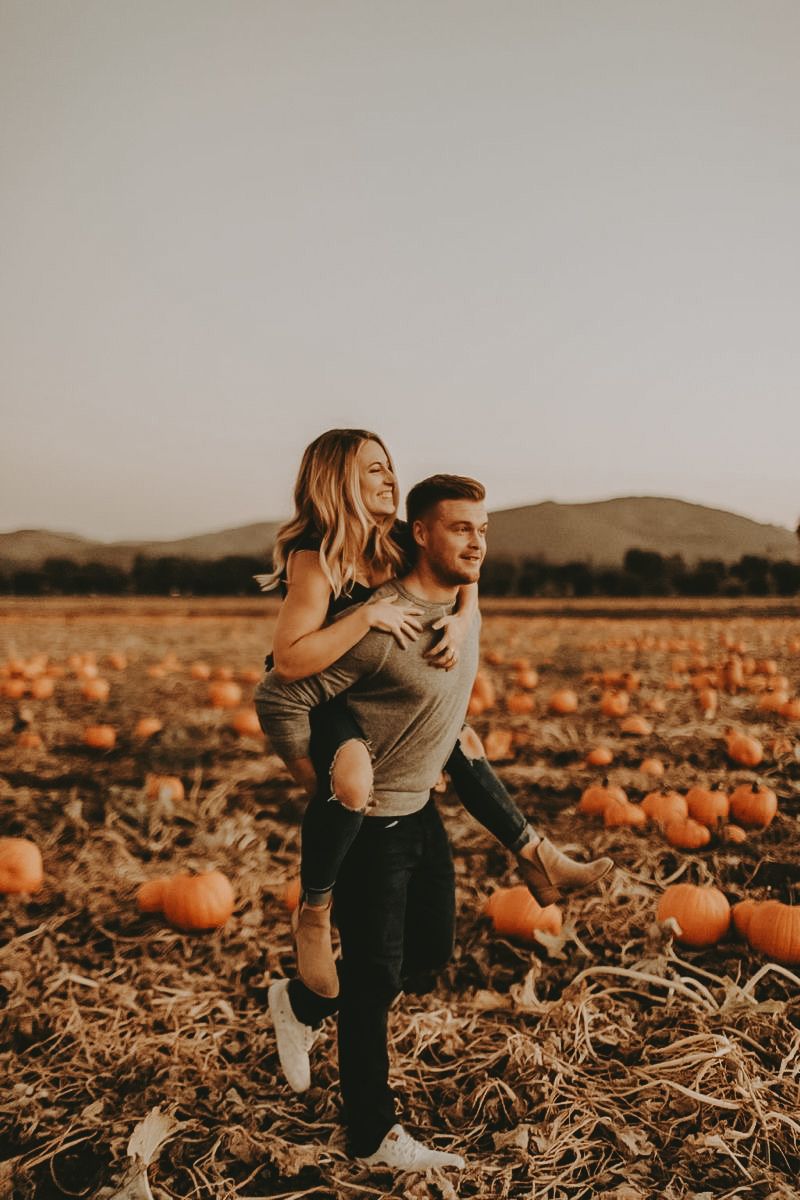 A couple enjoys a piggyback ride in a pumpkin patch field with rolling hills in the background.