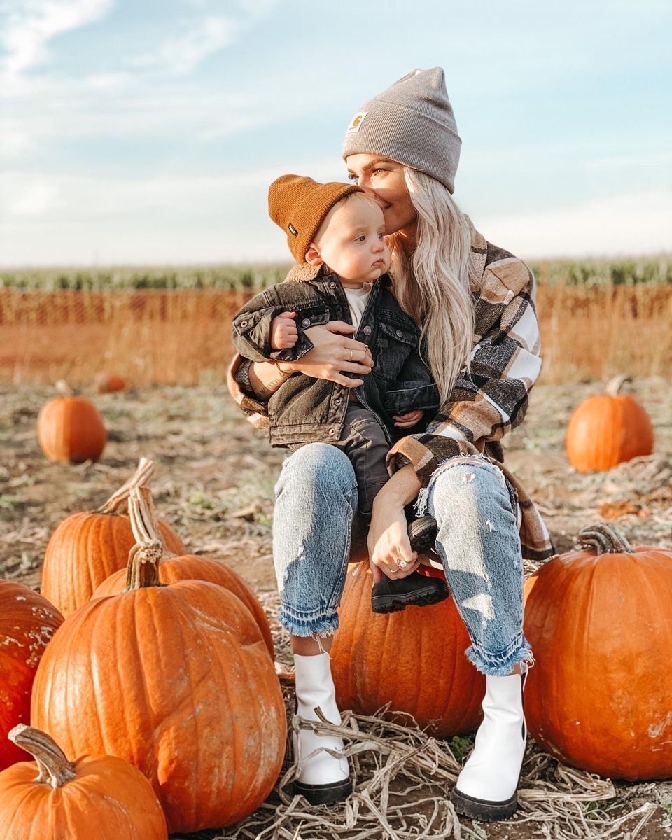 Mother kissing toddler at autumn pumpkin patch, both wearing cozy fall outfits with beanies, surrounded by orange pumpkins.