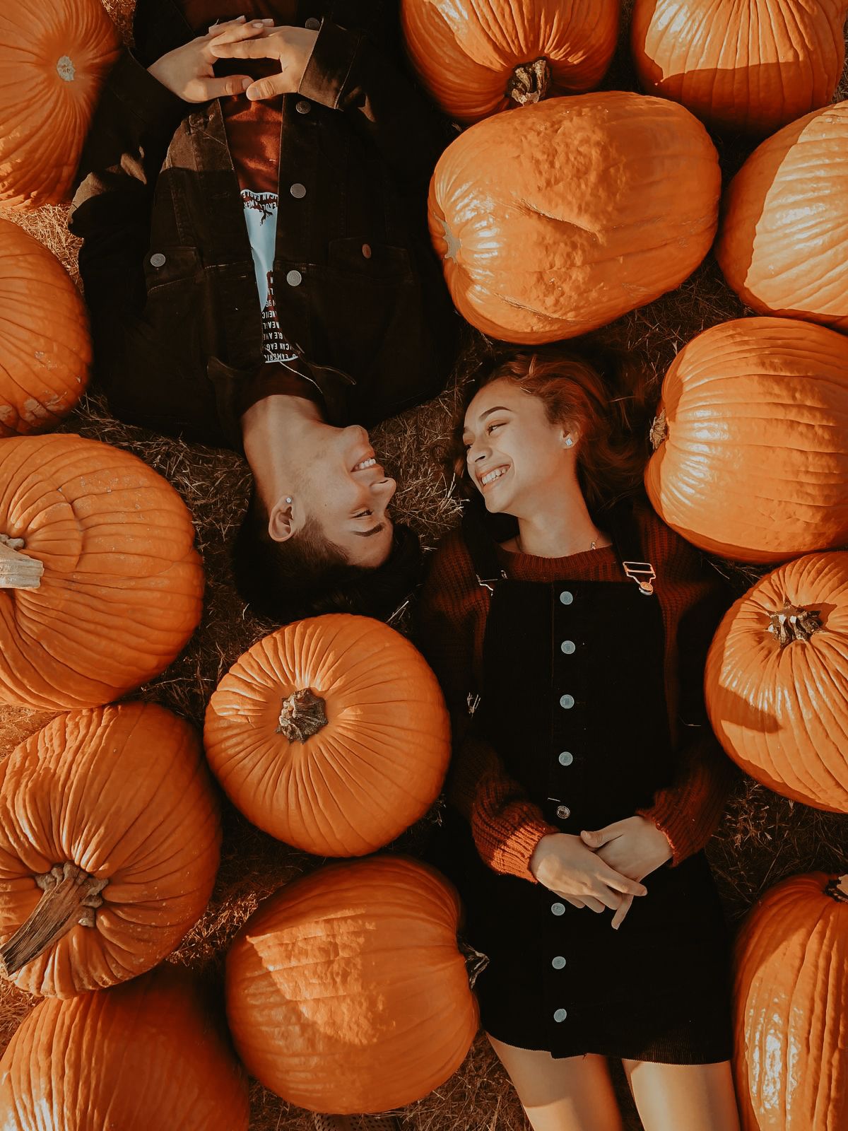 Couple lying among orange pumpkins in a pumpkin patch, smiling at each other in a romantic fall setting.