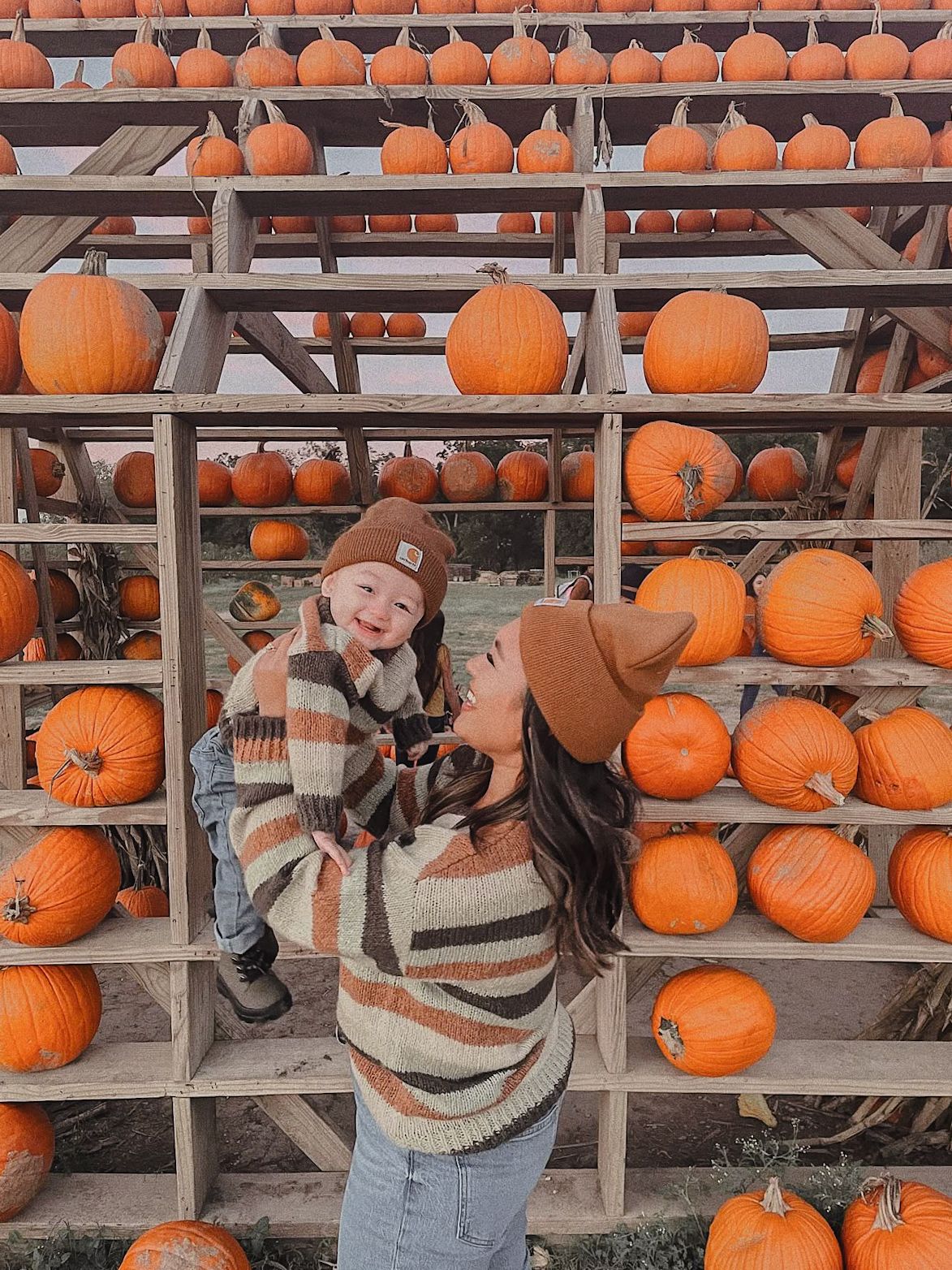 A mother holds her laughing baby at a fall pumpkin patch, both wearing cozy autumn sweaters and beanies.