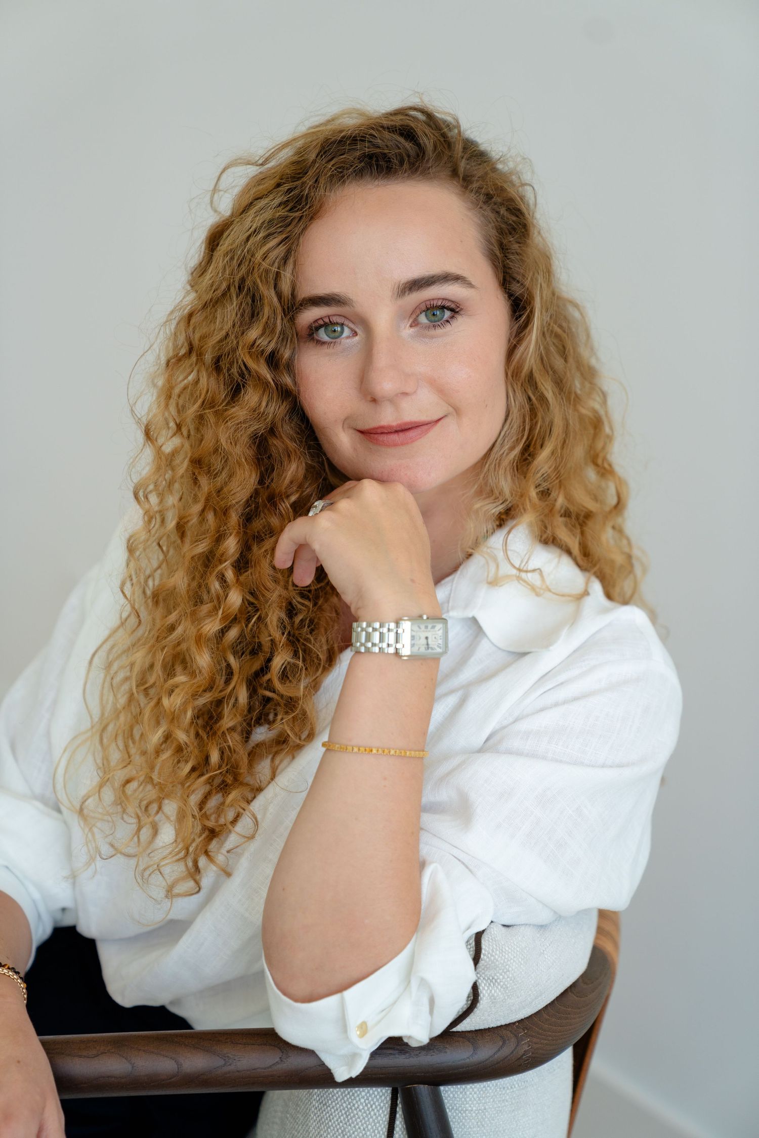 Young woman with curly blonde hair wearing a white shirt and silver watch, smiling while seated against a light background.