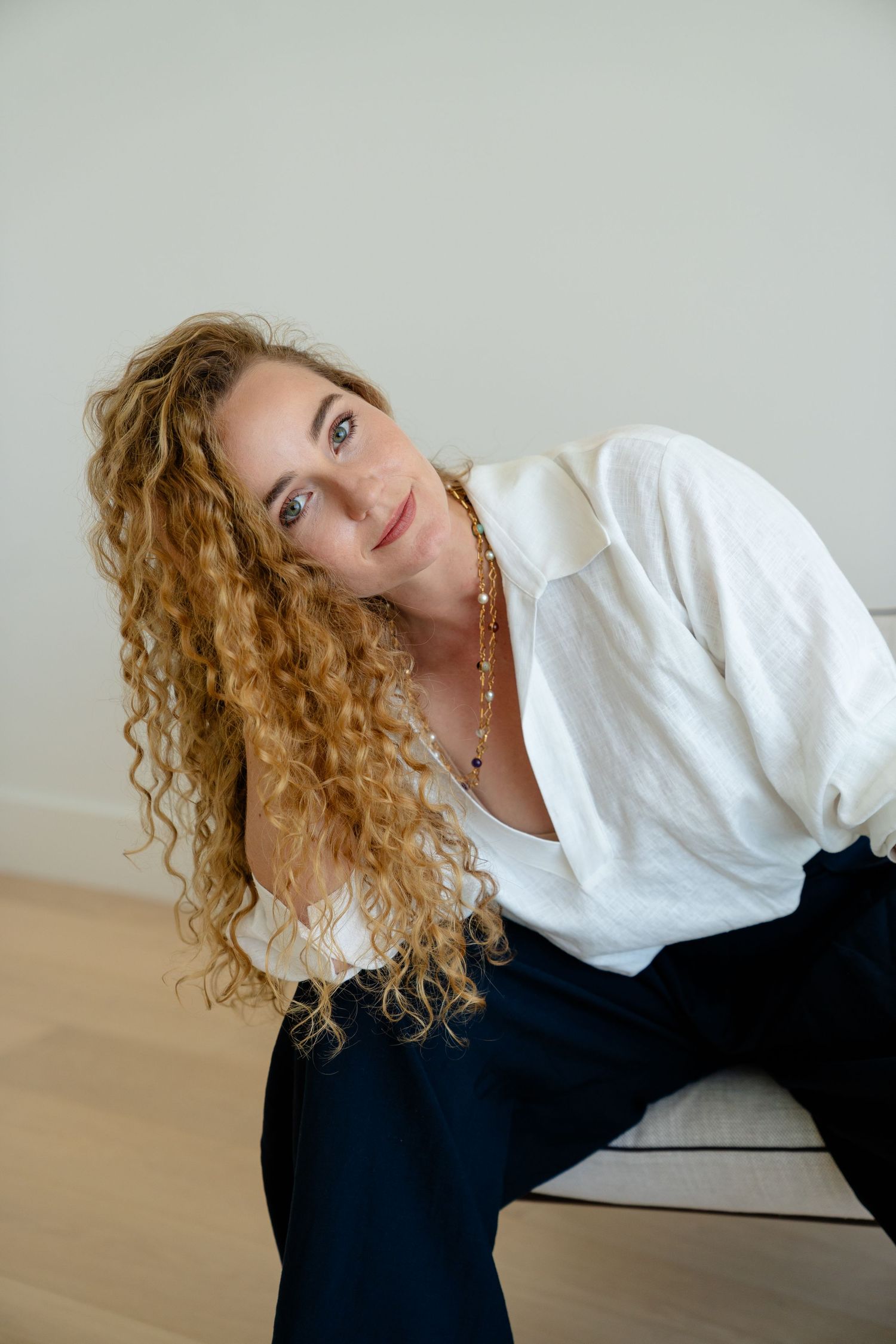 Woman with curly blonde hair in white shirt and navy pants poses gracefully on chair against neutral background.