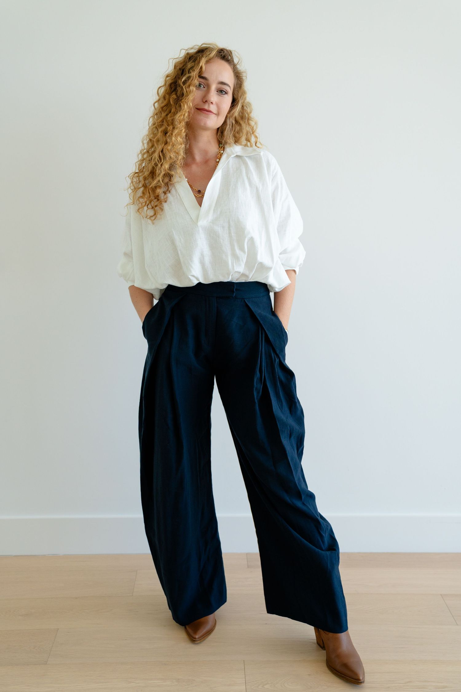 Woman in white oversized shirt and navy wide-leg trousers with brown ankle boots posing against white wall.