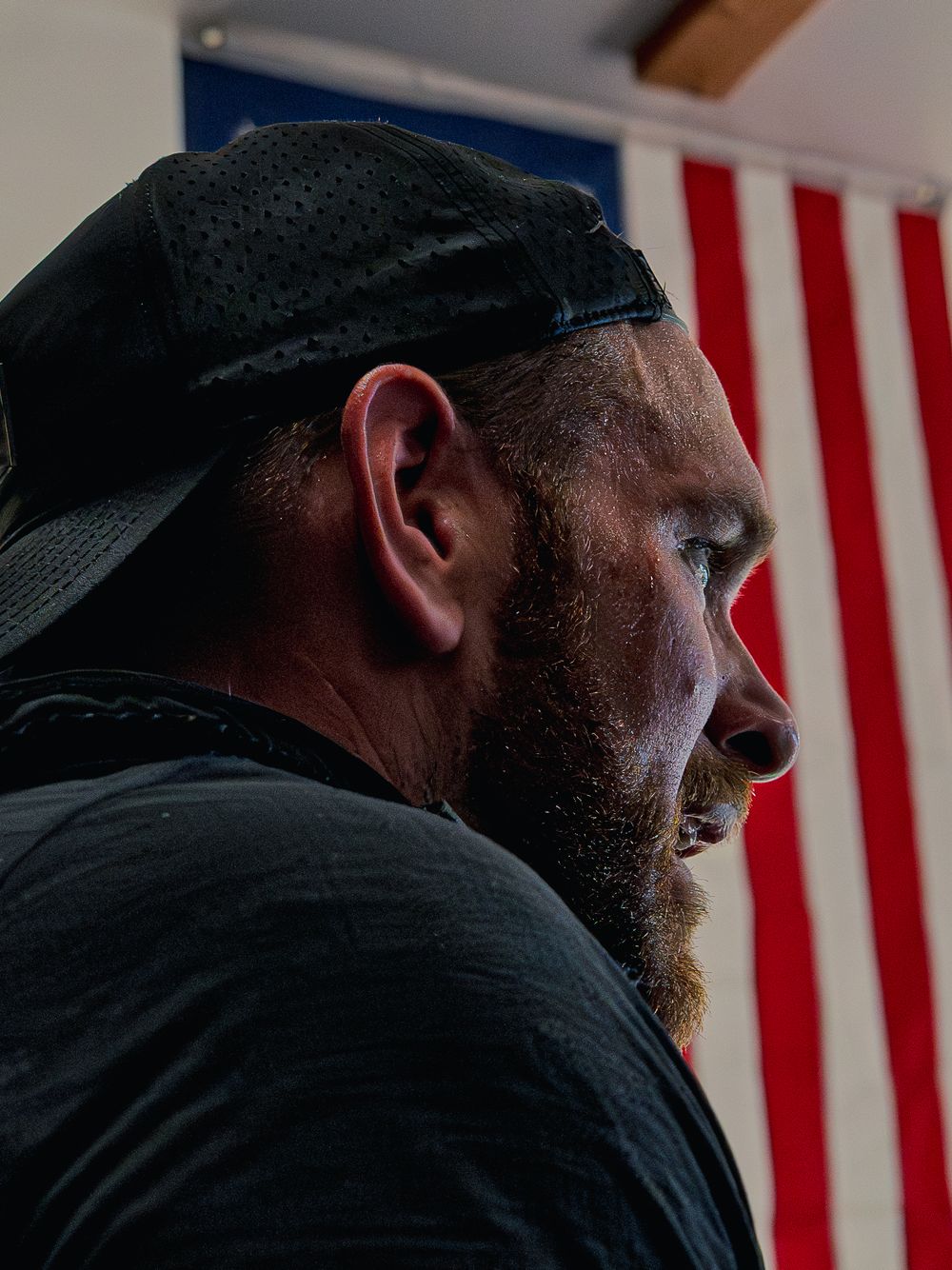 Close side profile of a sweat-drenched CrossFit athlete in a black cap catching his breath during Murph, an American flag behind him.