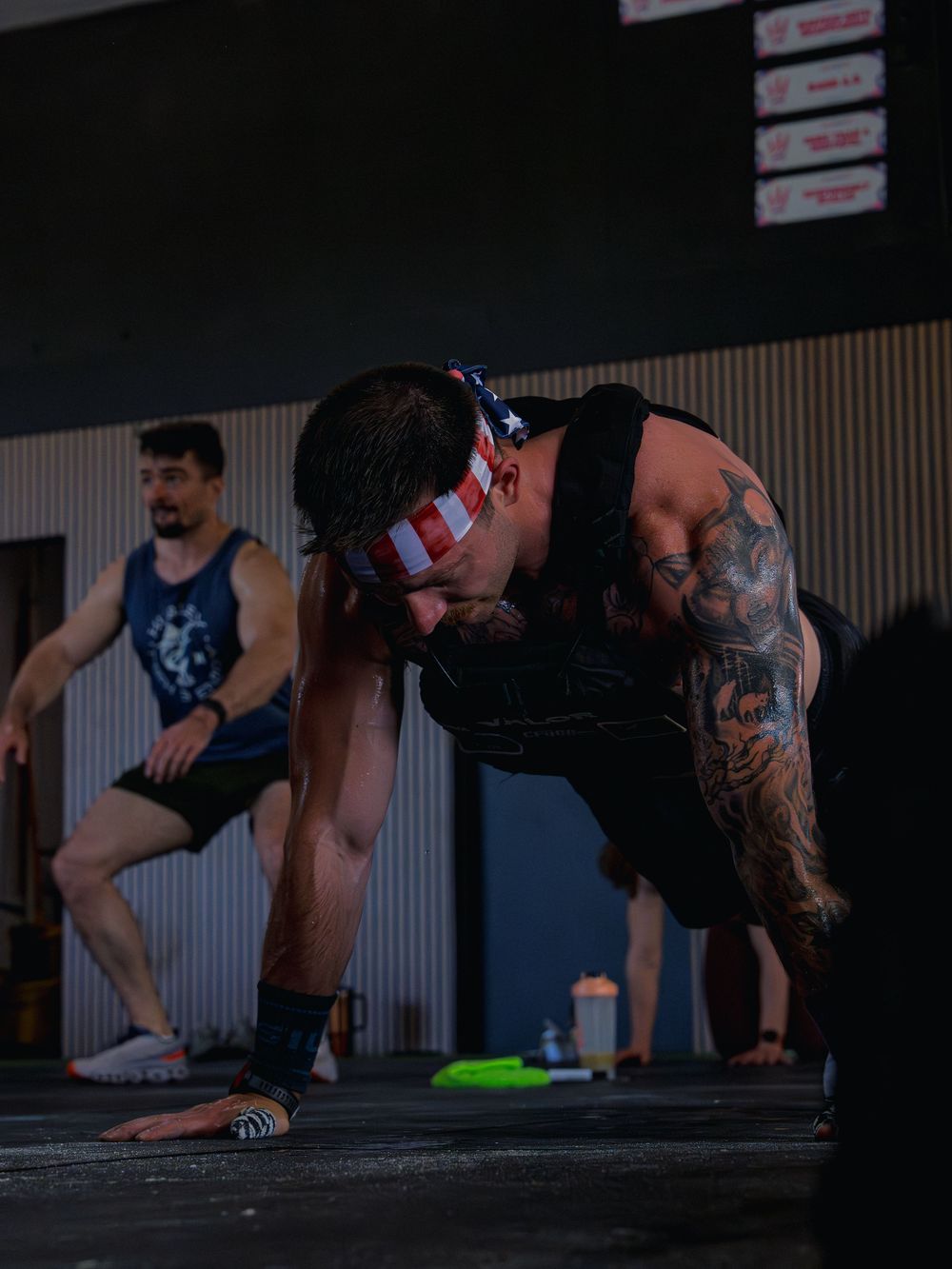 Tattooed CrossFit athlete in a weight vest and flag headband holds the top of a push-up during Murph as another athlete works behind him.