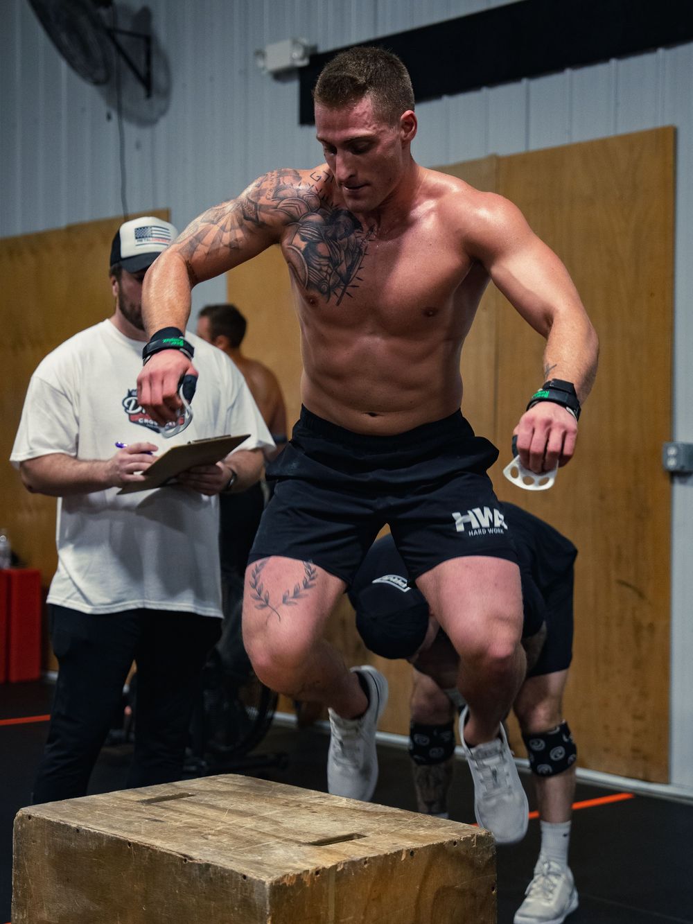 Tattooed CrossFit athlete leaps midair over a wooden box during a competition as a judge with a clipboard counts the rep behind him.
