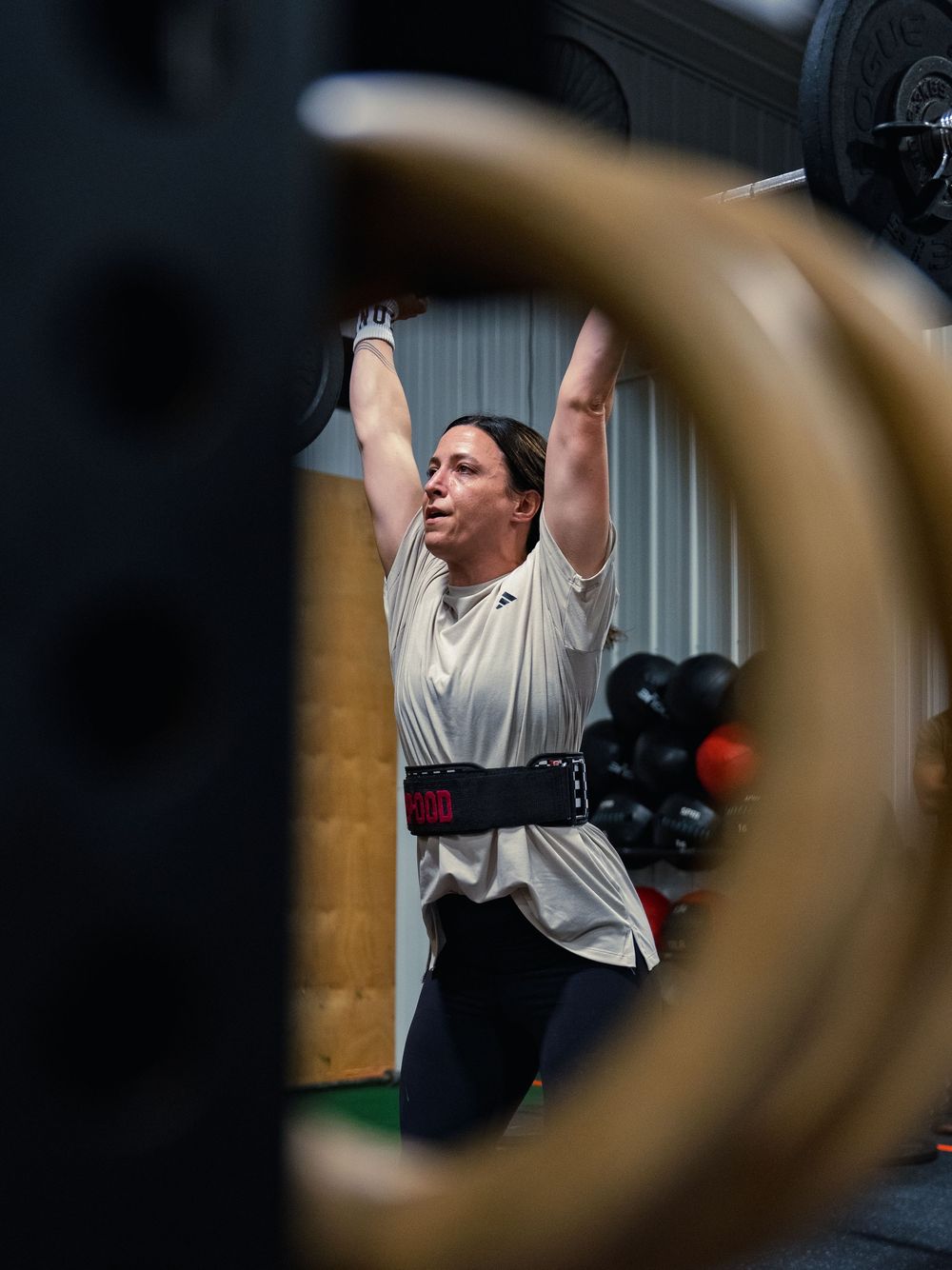 CrossFit athlete in a lifting belt locks a barbell overhead, framed through a bumper plate with medicine balls racked behind her.