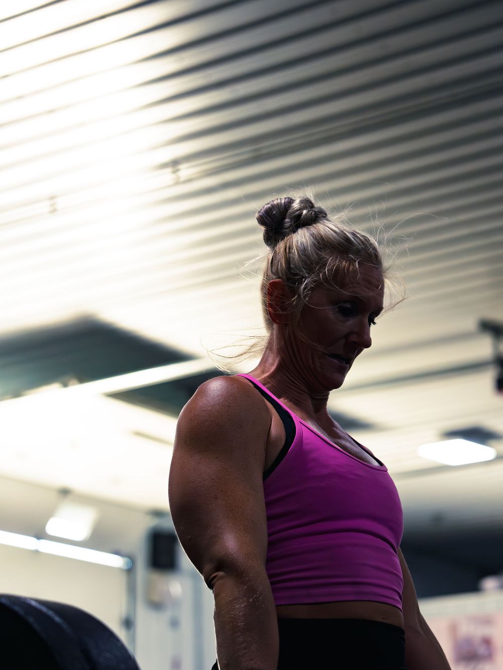 CrossFit athlete in a pink tank looks down to reset between sets, shoulders lit against the gym ceiling as she gathers herself mid-workout.