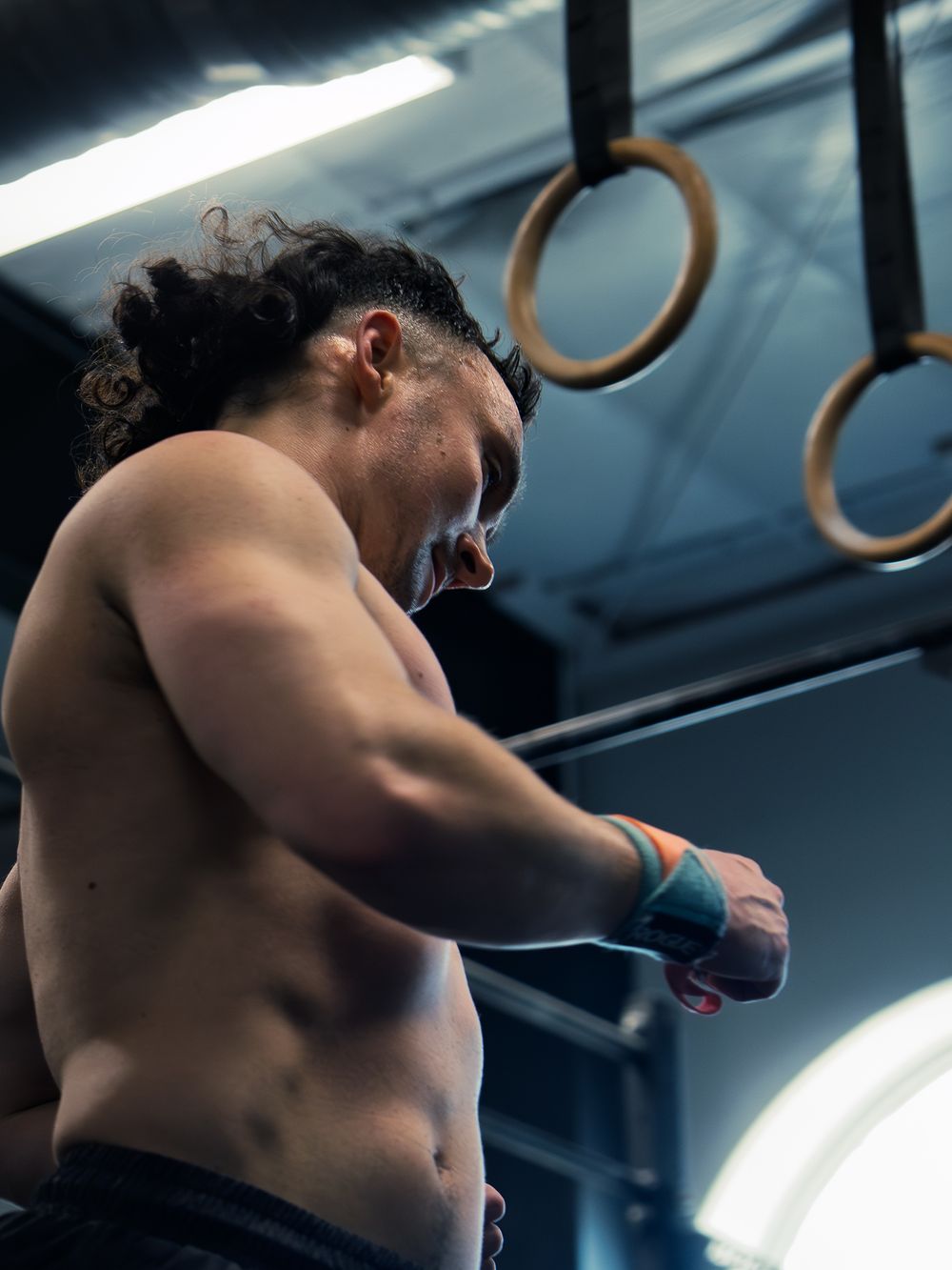CrossFit athlete in wrist wraps looks down to reset between rounds, gymnastics rings hanging above him as he catches his breath mid-workout.