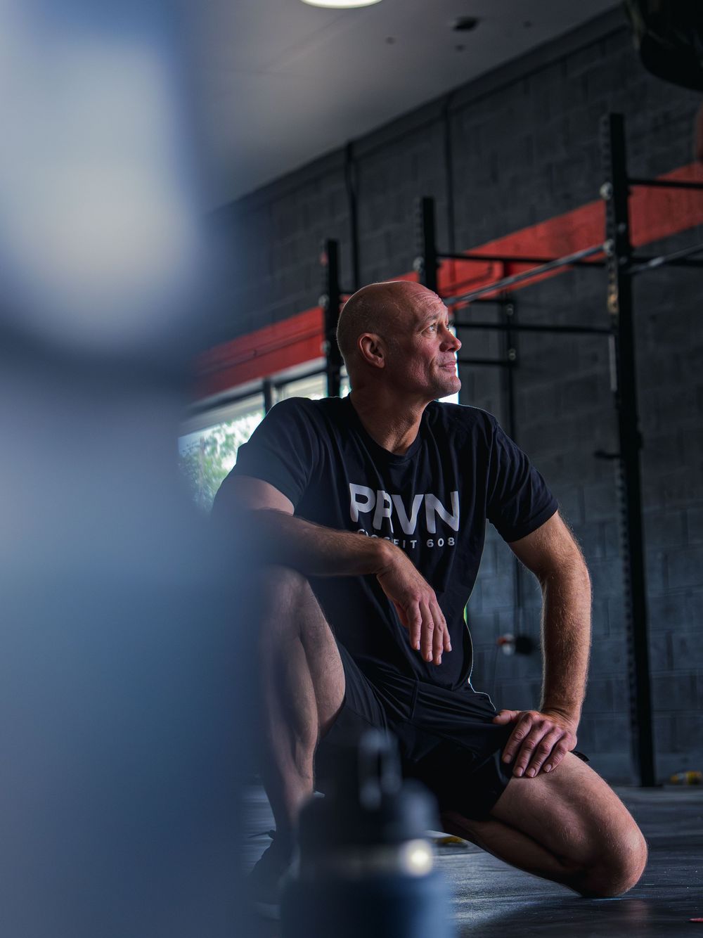 CrossFit 608 athlete in a PRVN tee kneels to rest between rounds, gaze fixed toward the window light as he catches his breath mid-workout.