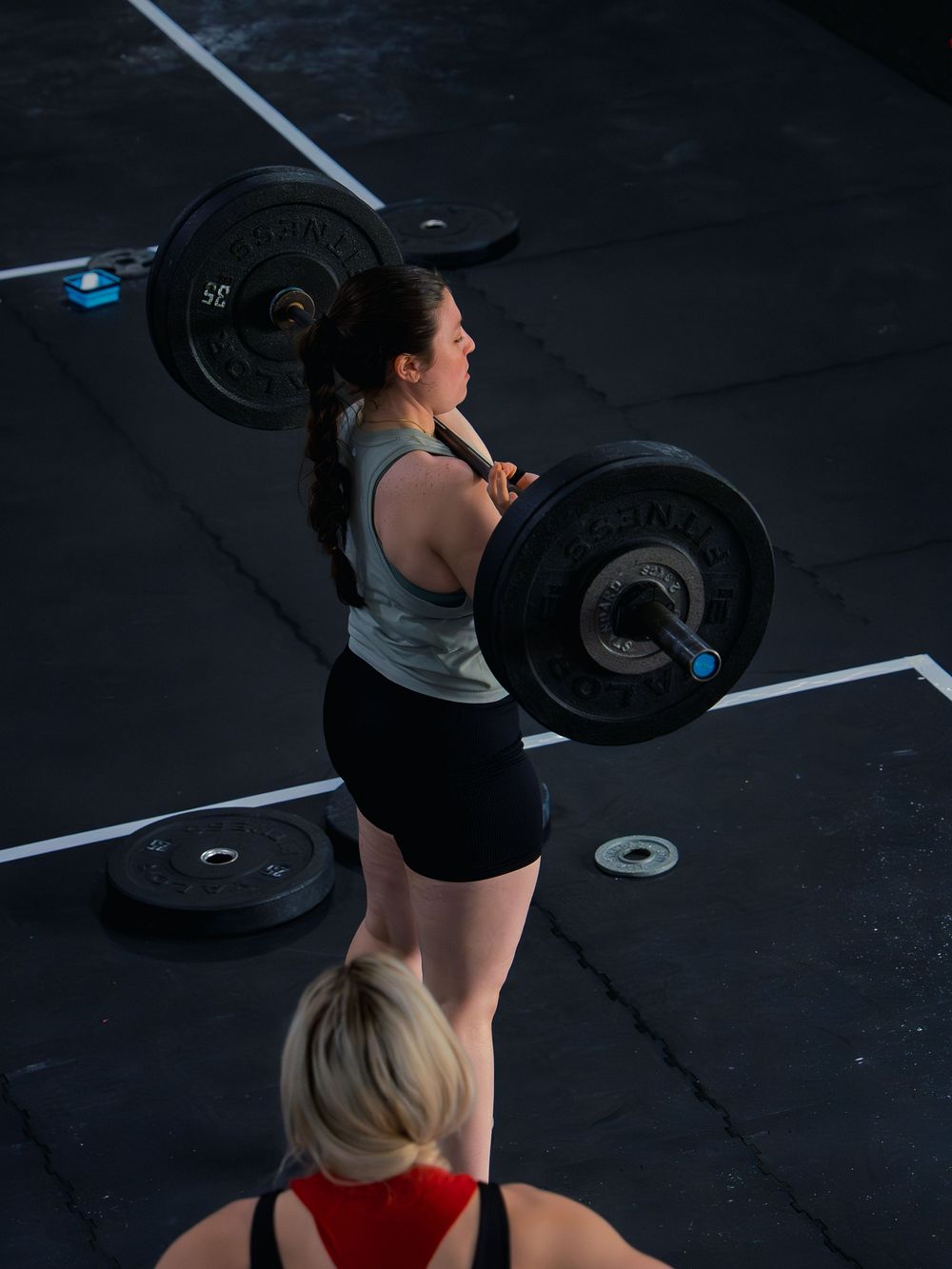 CrossFit athlete holds a loaded barbell in the front rack, shot from above as a judge watches from below during a competition workout.