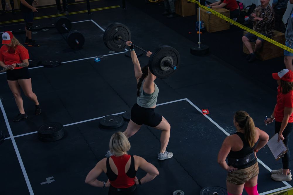 CrossFit athlete locks a loaded barbell overhead in a split jerk at a competition in Janesville, WI, judges tracking the rep close by.