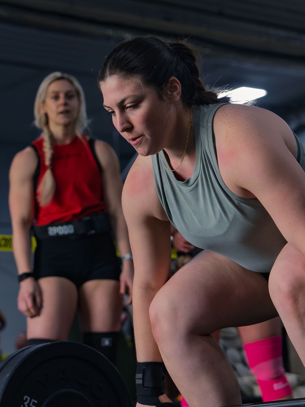 CrossFit athlete sets her grip and braces over a loaded barbell before a lift at a competition, a judge watching close behind her.