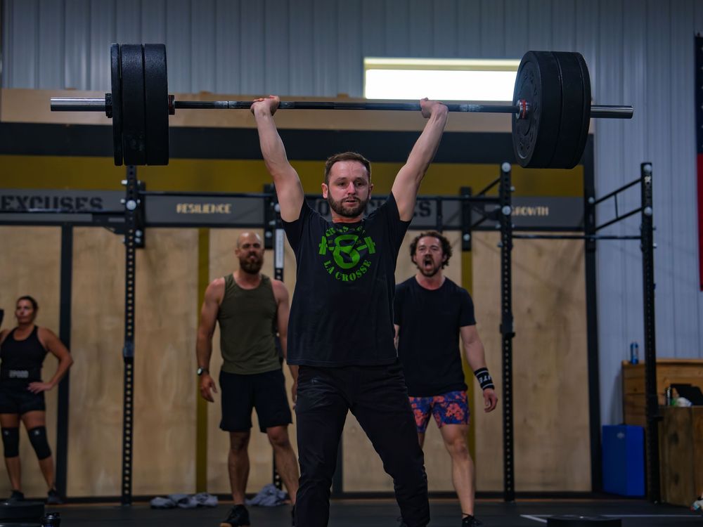 CrossFit athlete locks a heavy loaded barbell overhead in a jerk at a competition in Janesville, WI, teammates cheering him on behind.