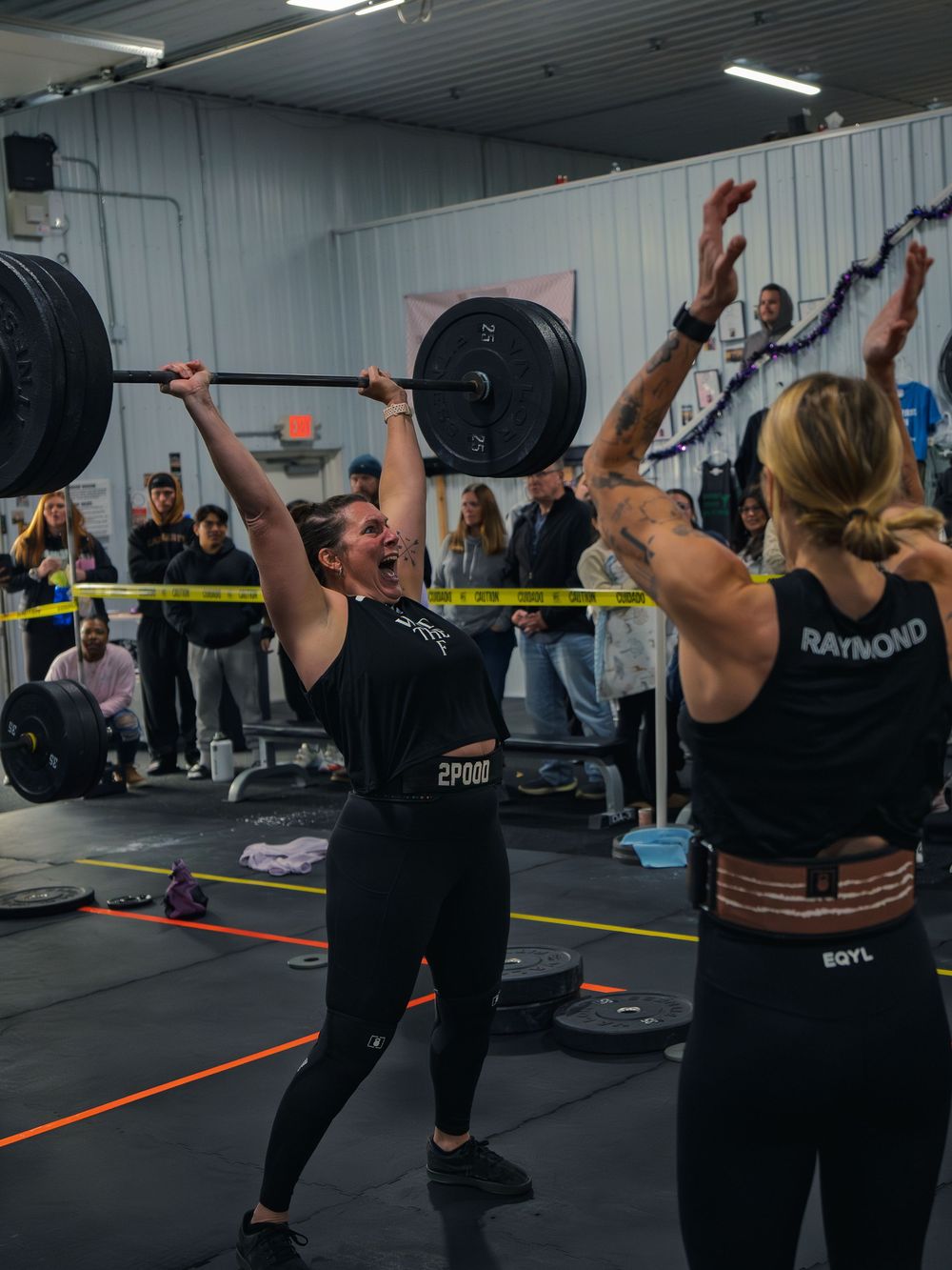 CrossFit athlete grinds a loaded barbell to lockout overhead, face straining, as a judge counts reps and the crowd watches at a competition.
