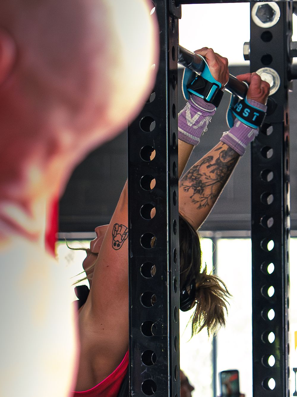 CrossFit athlete in gymnastics grips hangs from the pull-up bar between reps, tattooed arms extended and head tipped back through the rig.