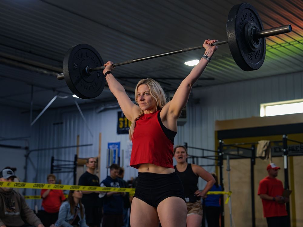 CrossFit athlete in a red tank locks a loaded barbell overhead in a snatch at a competition, arms fully extended as the crowd looks on.