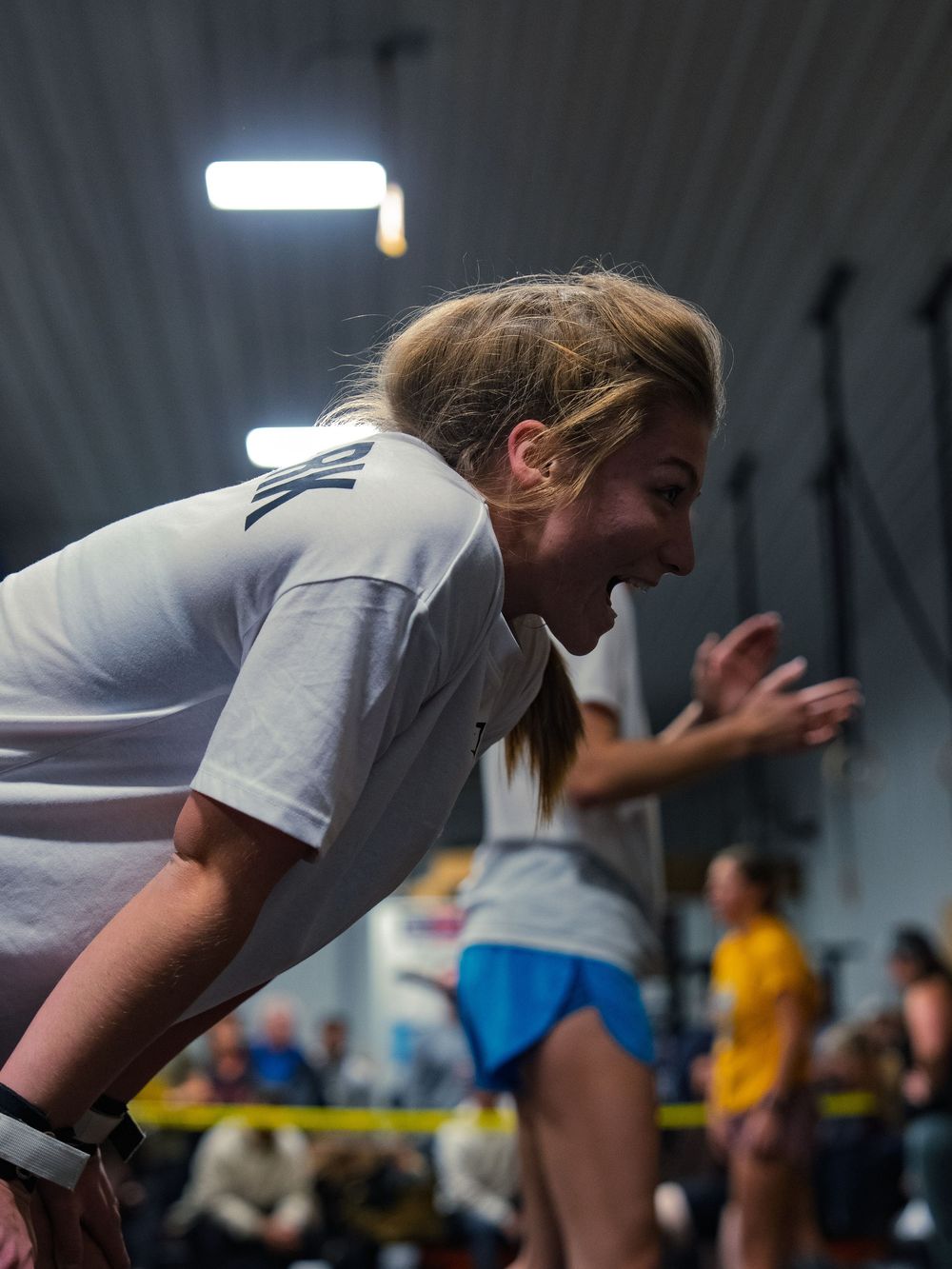 CrossFit athlete on the sideline shouts encouragement to a teammate mid-workout at a competition, face lit up as another cheers behind her.