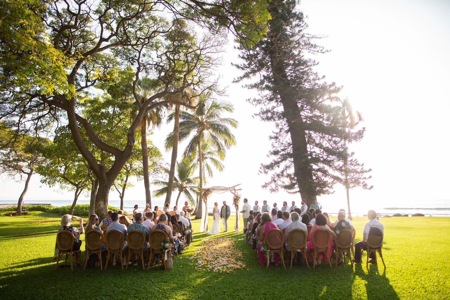 Bride and groom kissing on a rocky Hawaiian beach at sunset with ocean and mountain views.