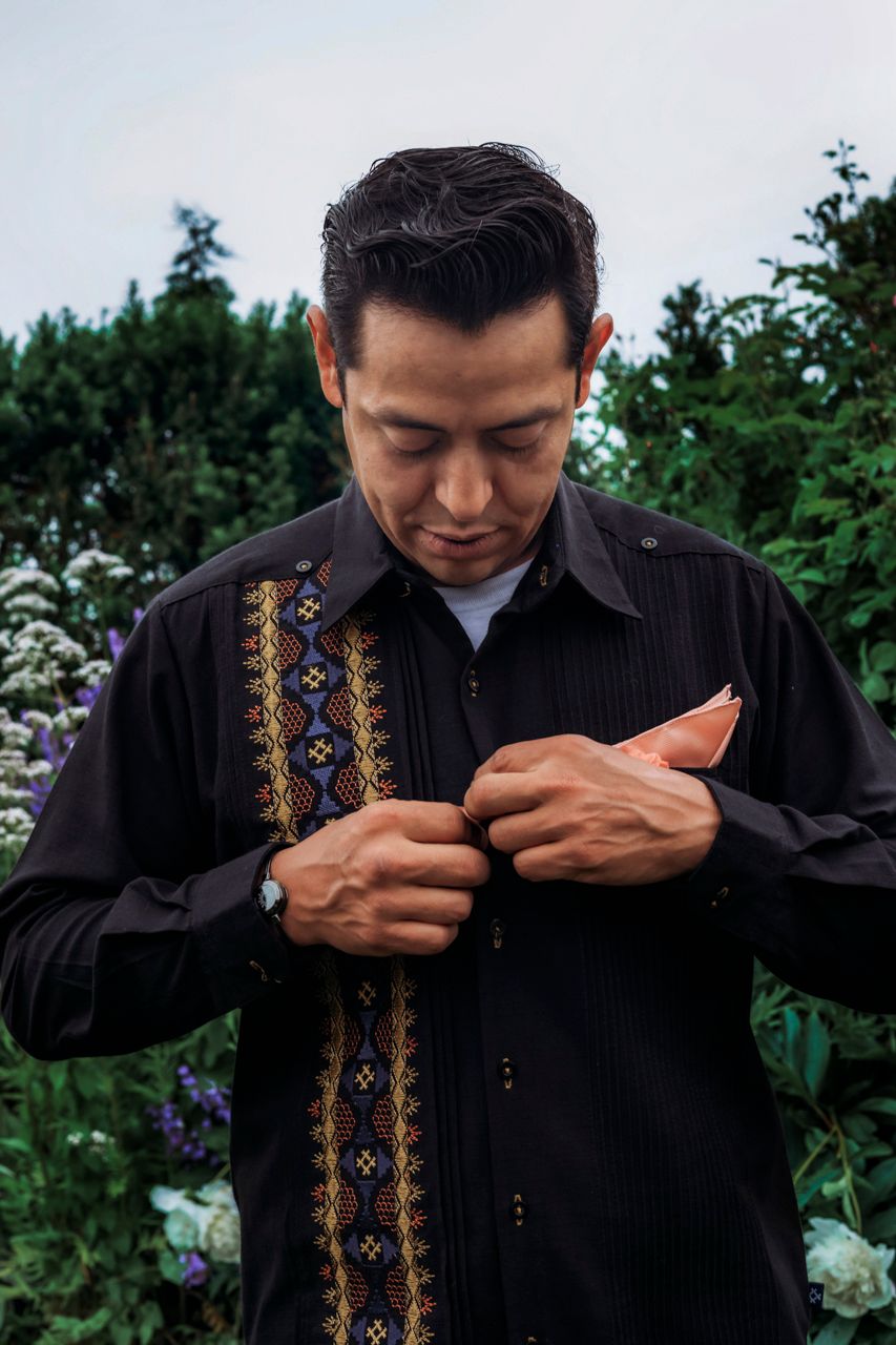 Man in black embroidered guayabera shirt buttoning up outdoors with green trees and flowers in background.
