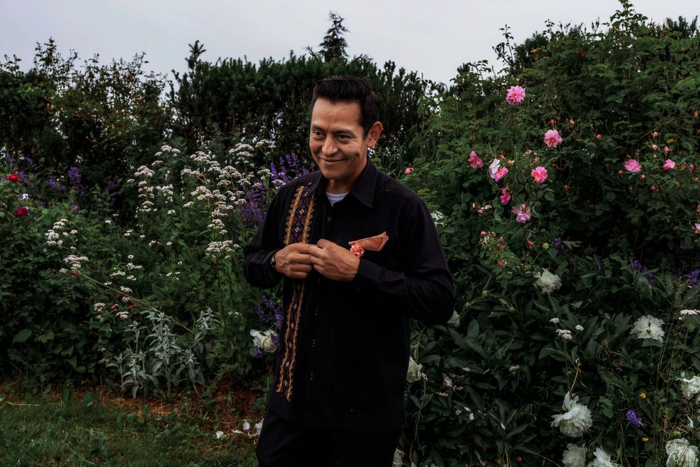 Man in black outfit smiling in a lush garden with colorful roses and white flowers blooming in the background.
