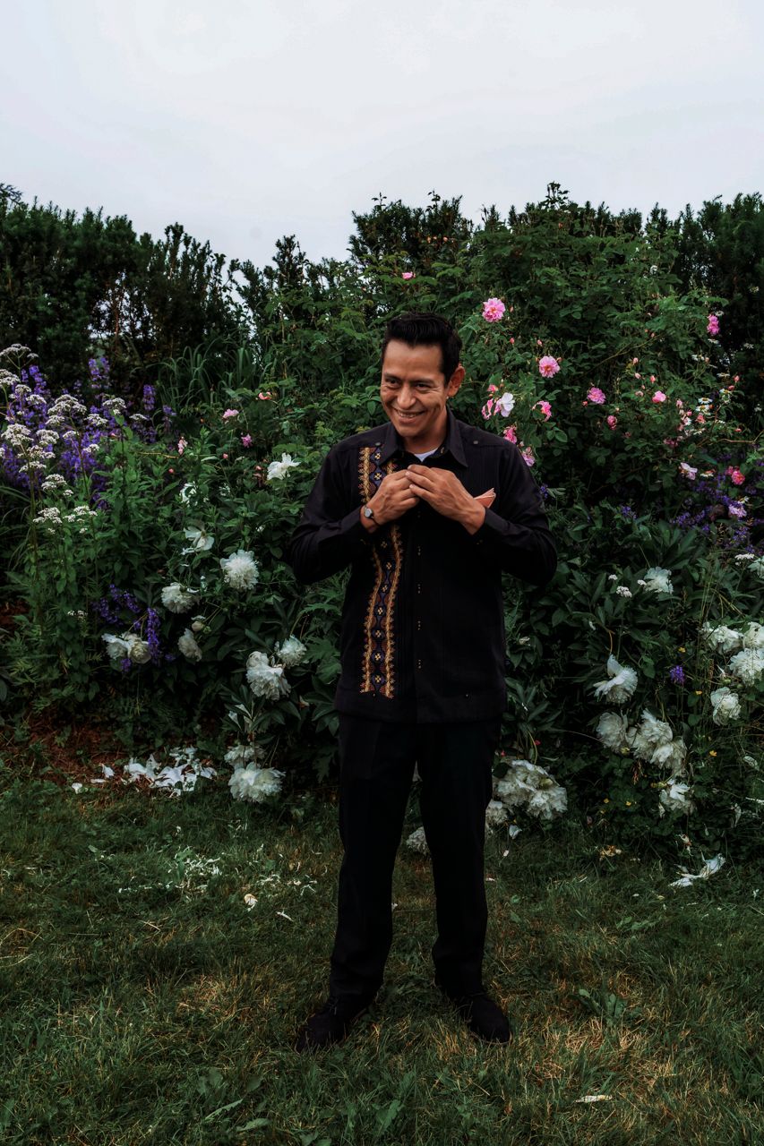Man in black outfit smiling in a lush garden with colorful flowers including roses and purple blooms.