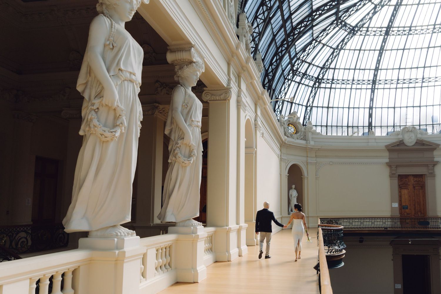 A couple walks hand-in-hand through a grand museum hall with classical caryatid statues and glass dome.