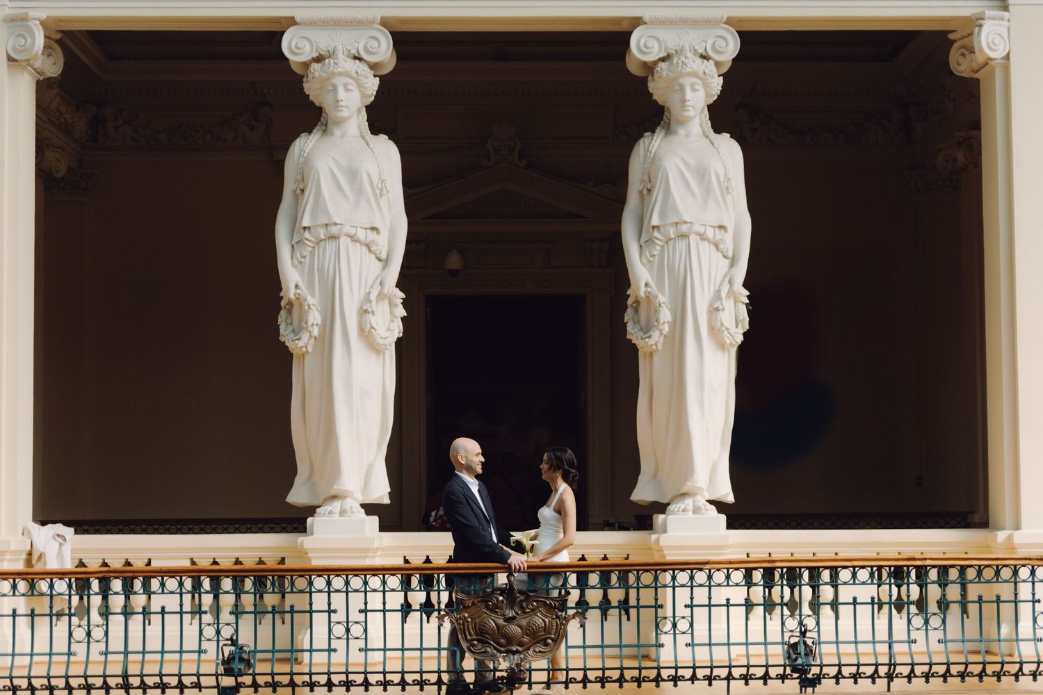 A wedding couple holds hands on a decorative balcony flanked by two white classical caryatid statues.