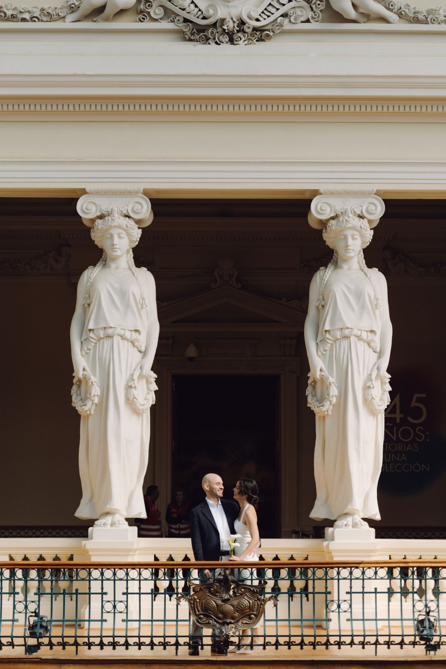 A wedding couple stands between two elegant caryatid statues on an ornate classical building balcony.