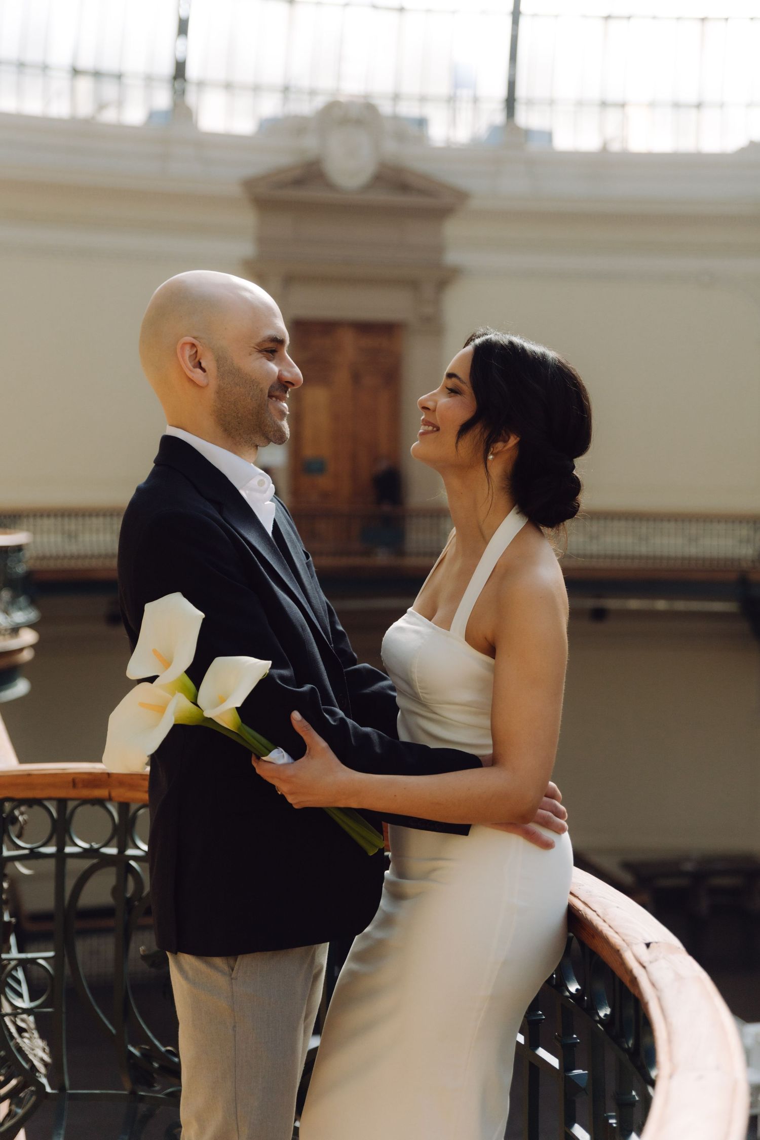 A laughing bride in white halter dress embraces a smiling bald groom holding calla lily bouquet indoors.