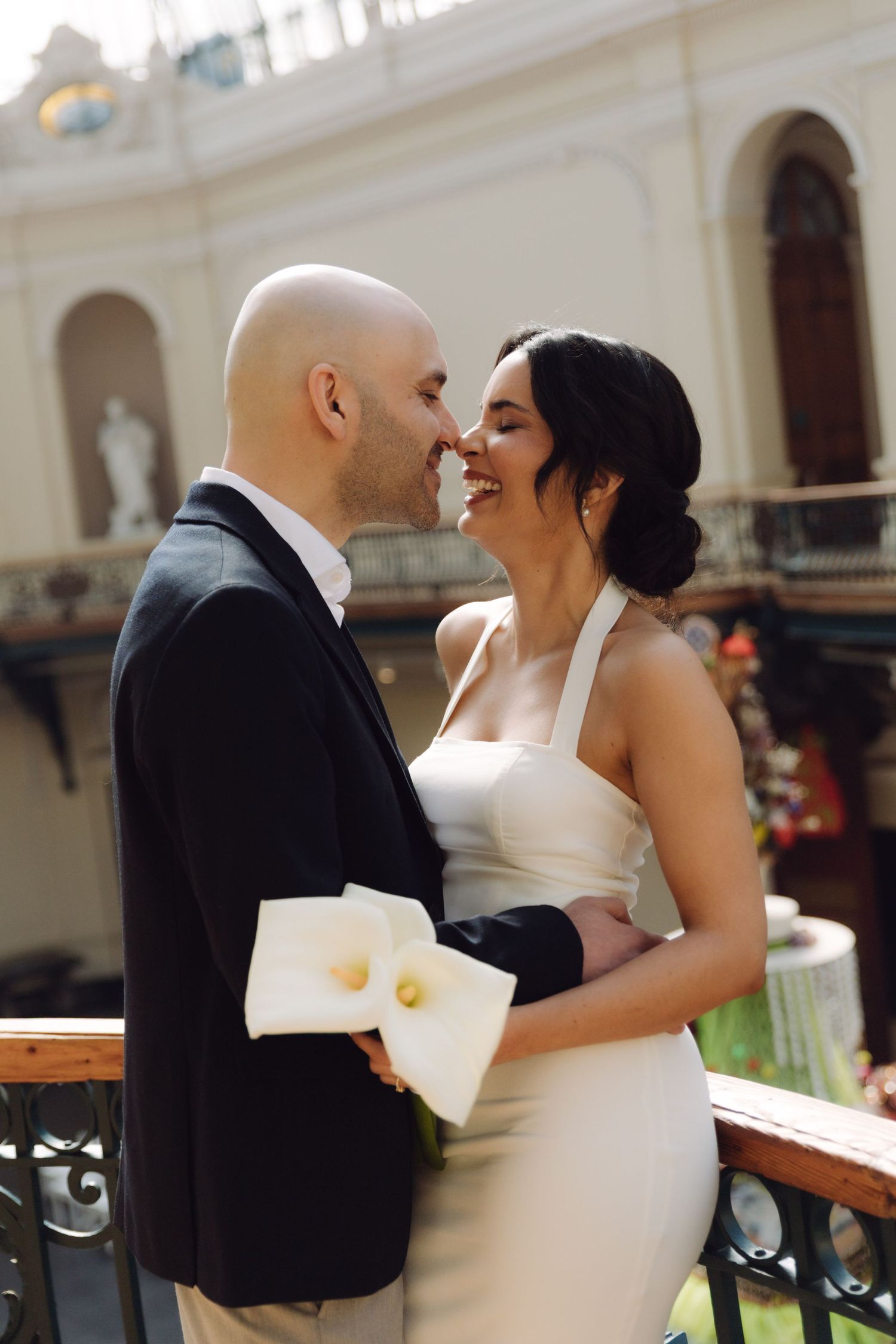 A bride and groom lean in for a kiss on a balcony inside a grand classical architectural venue.
