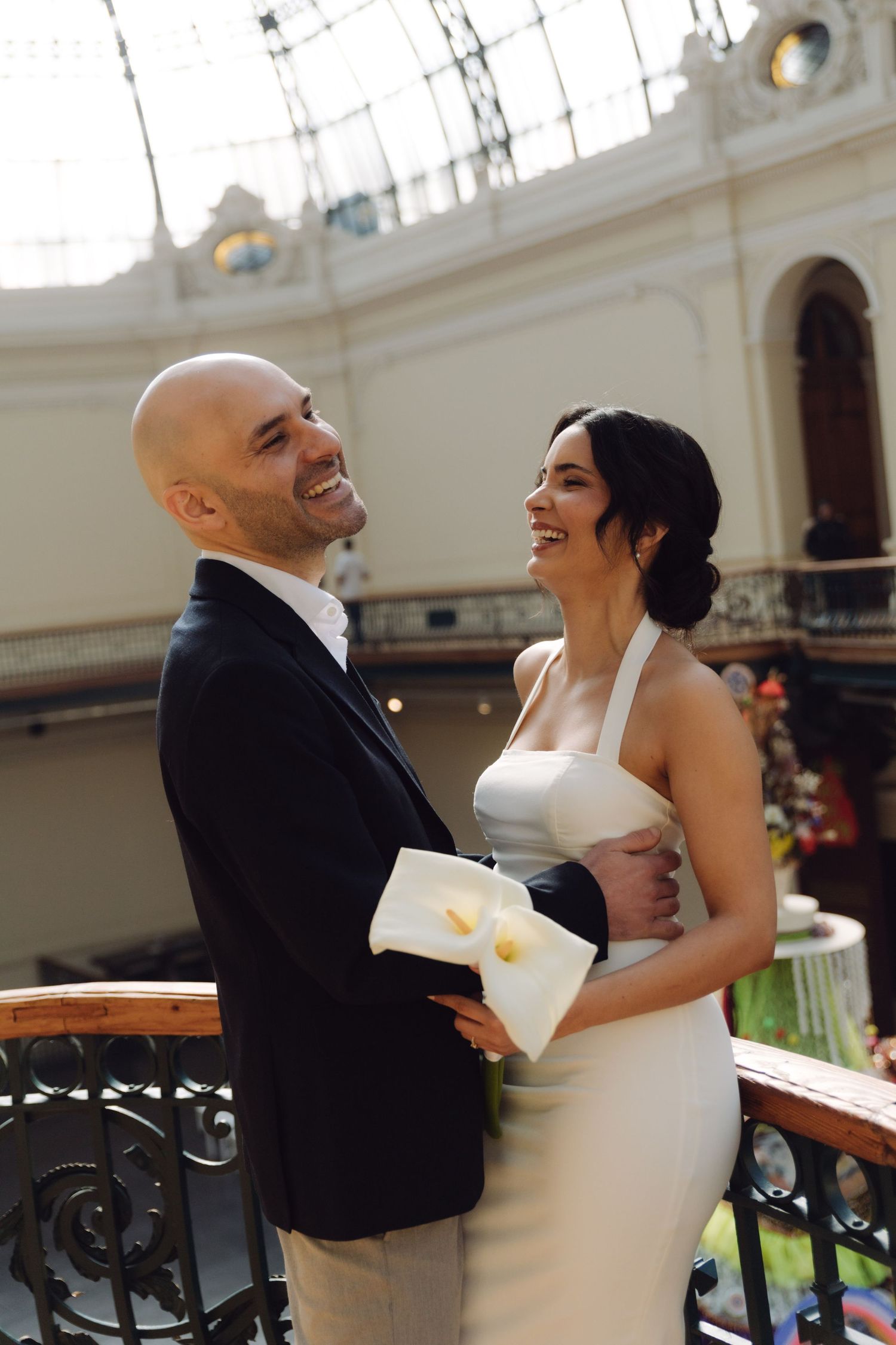 A joyful bride and bald groom laugh together on a balcony inside an elegant building with glass ceiling.