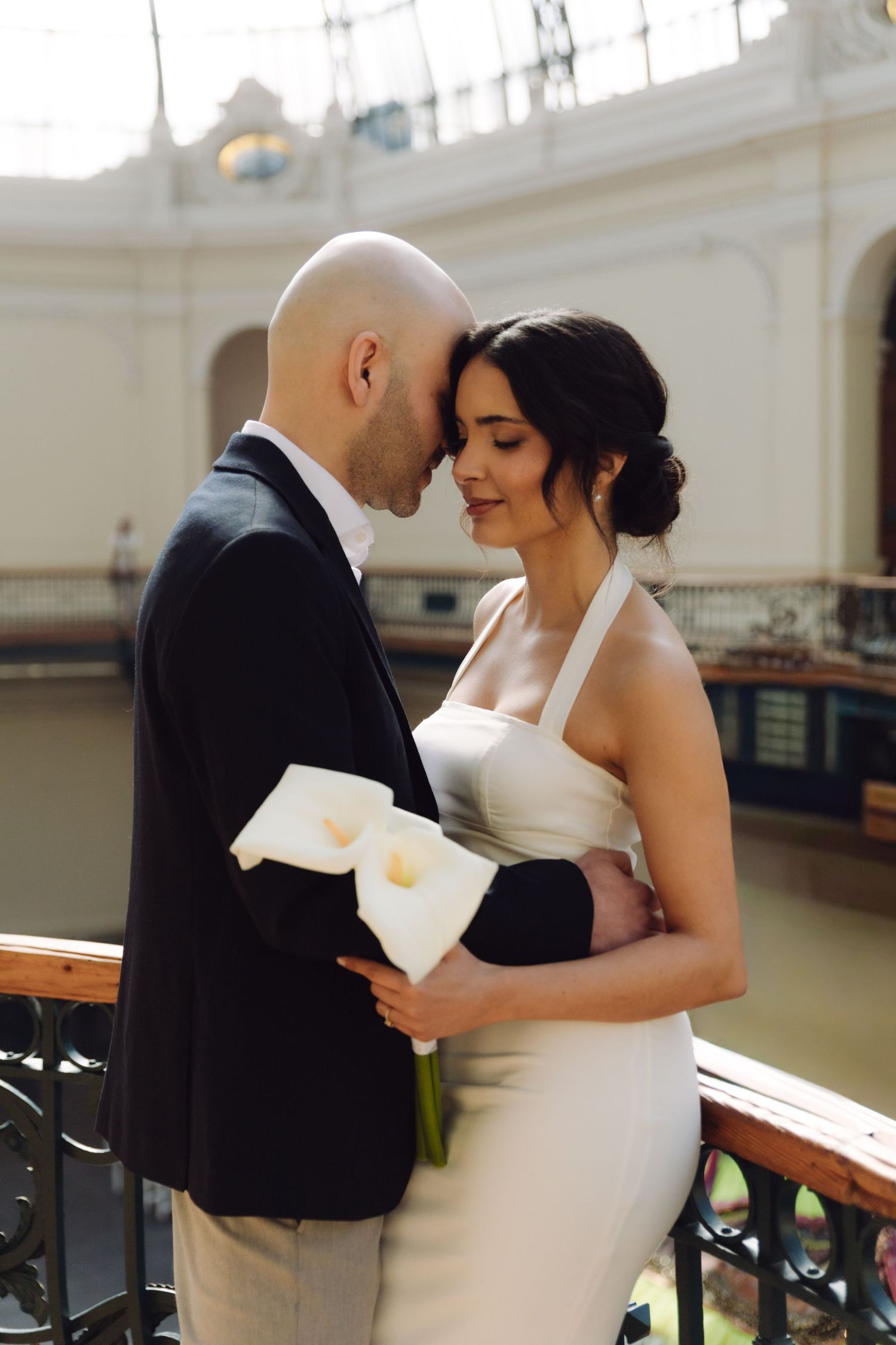 A bride and groom share a tender forehead touch while embracing on a balcony in a grand historic venue.