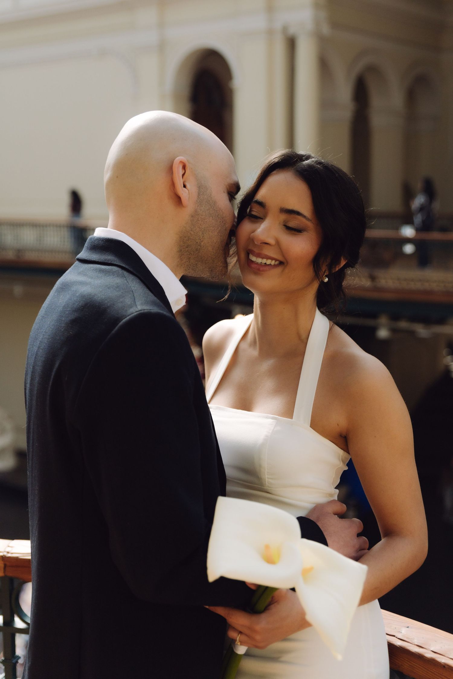A bald groom kisses his smiling bride on the cheek as she holds white calla lilies in a classic venue.
