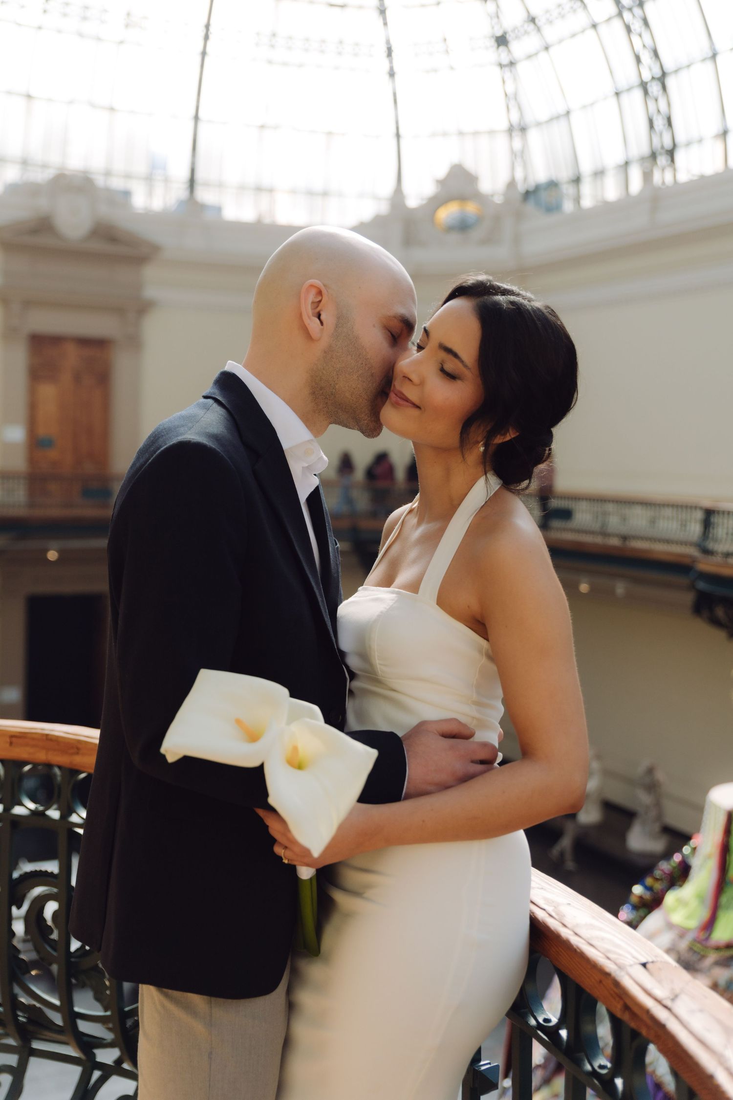 Bride and groom share a tender kiss on a balcony inside an elegant glass-domed venue, bride holding white calla lilies.