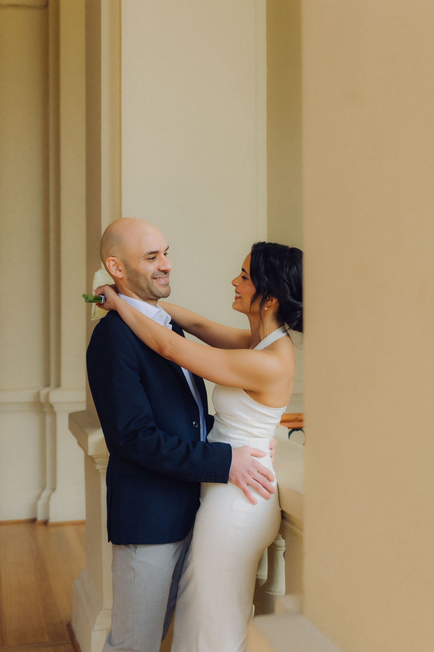 Smiling bride and groom embrace near classical columns, bride in sleek white halter gown, groom in navy blazer.