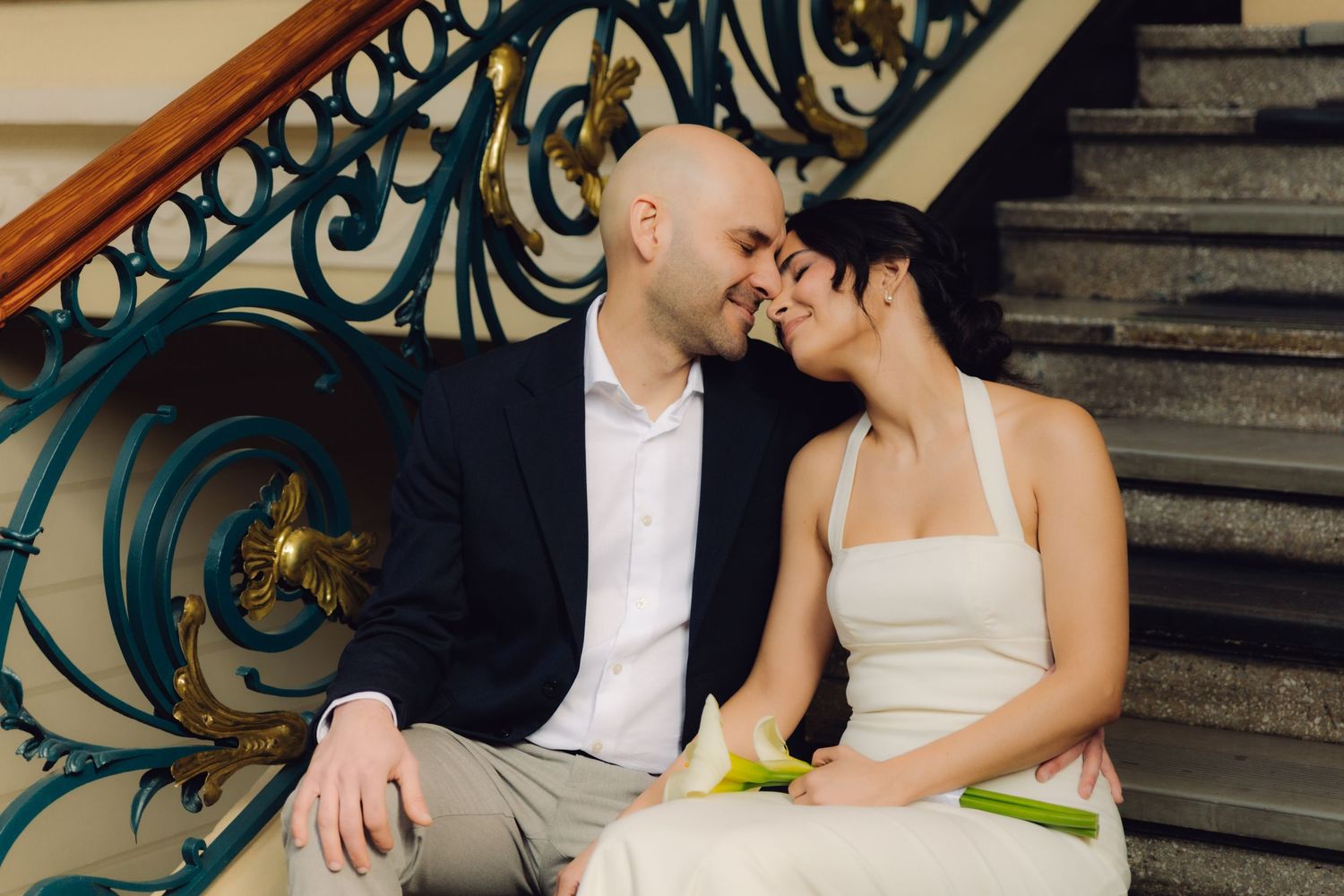 Bride and groom sit closely on ornate staircase steps, sharing an intimate moment with decorative iron railings.