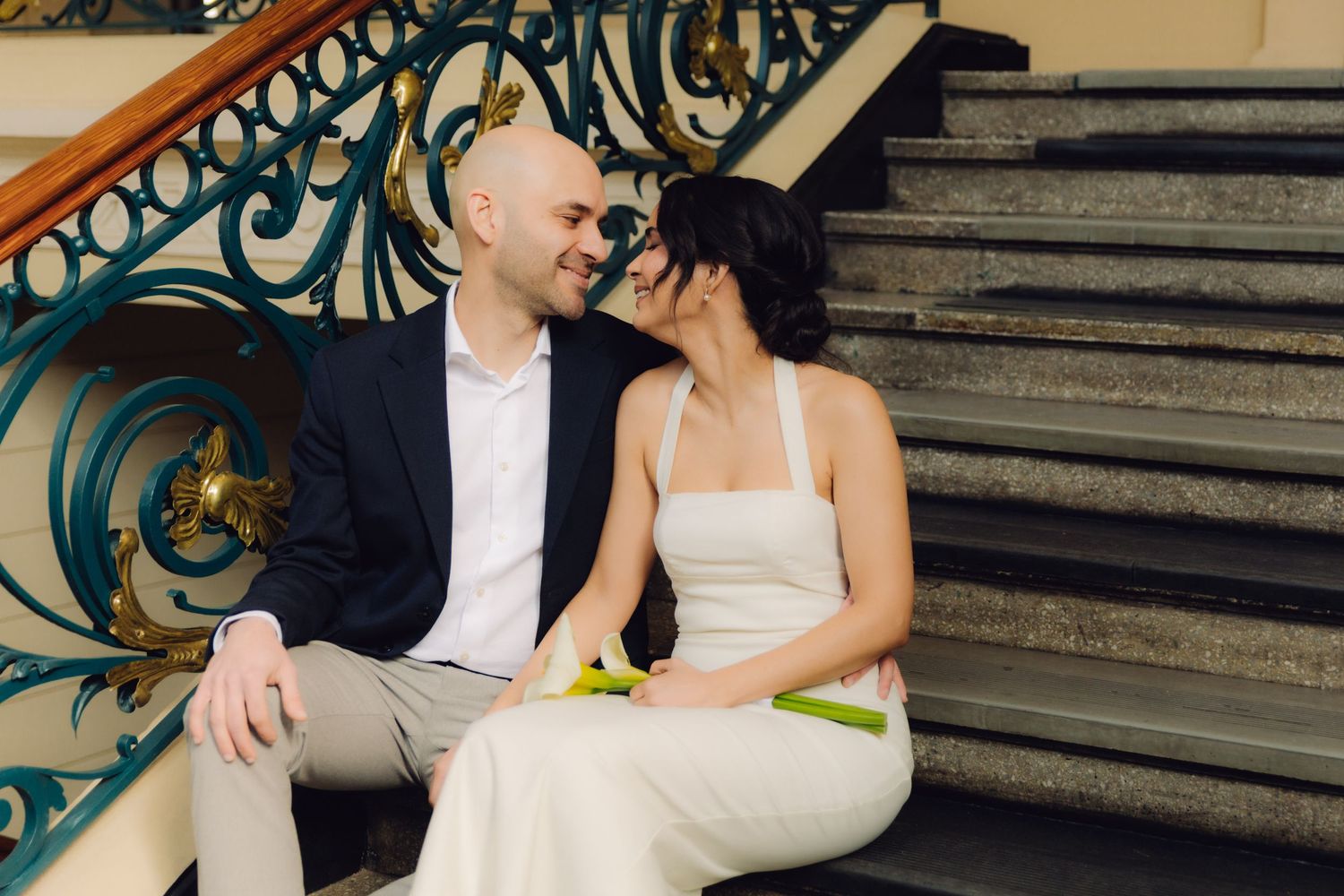 Bride and groom smile at each other while seated on grand staircase with elegant blue wrought iron balustrade.