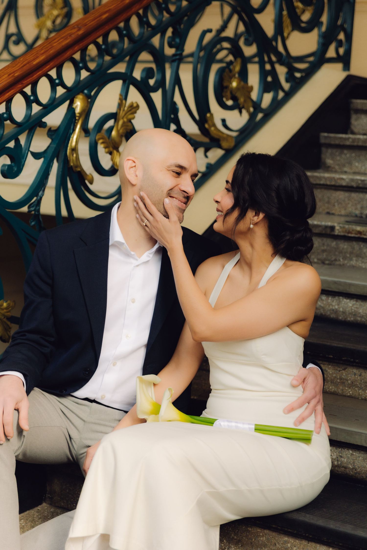 Bride tenderly touches grooms face while seated together on ornate staircase steps in a grand historic building.