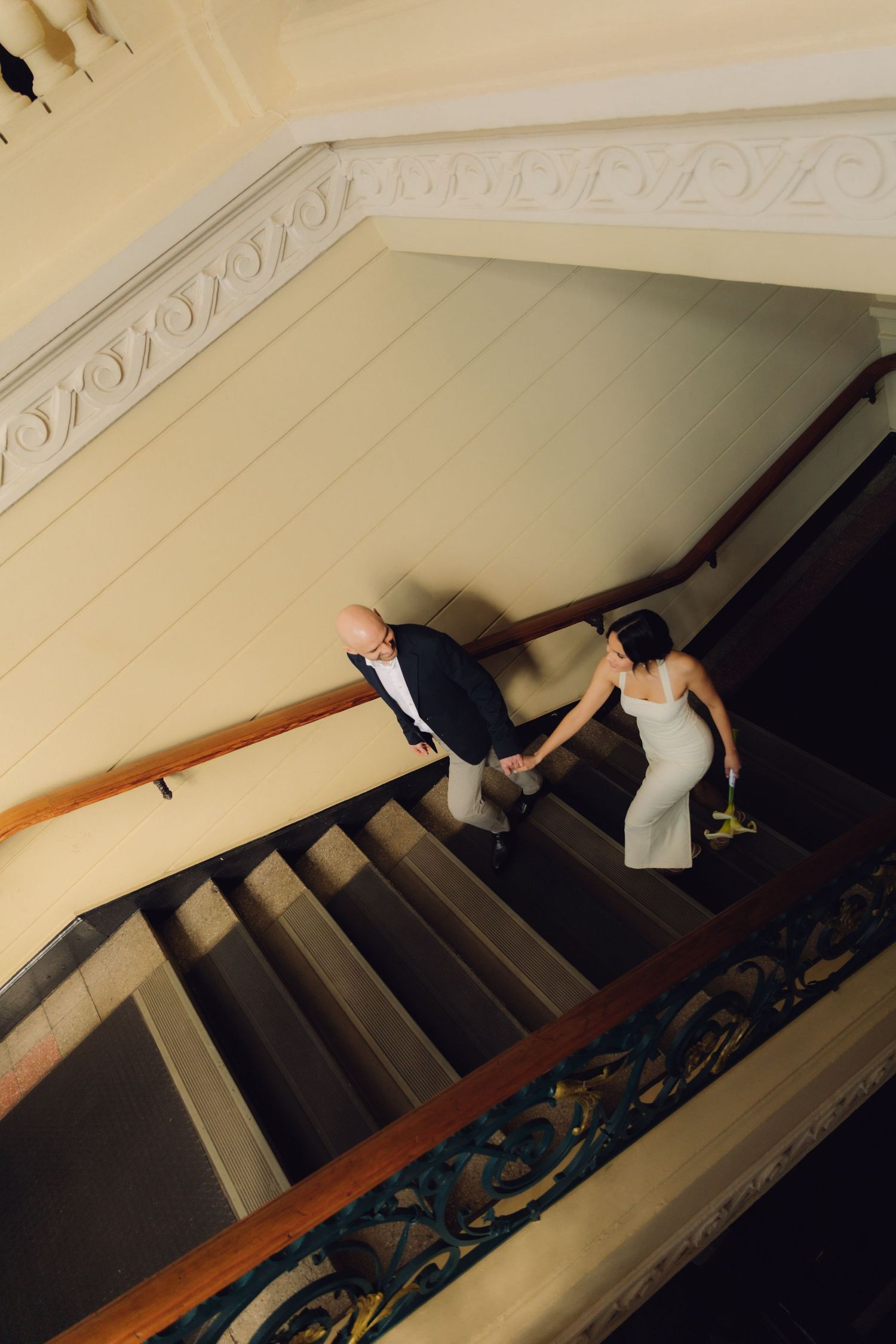 Aerial view of bride and groom holding hands while ascending a classic wooden staircase in an elegant venue.