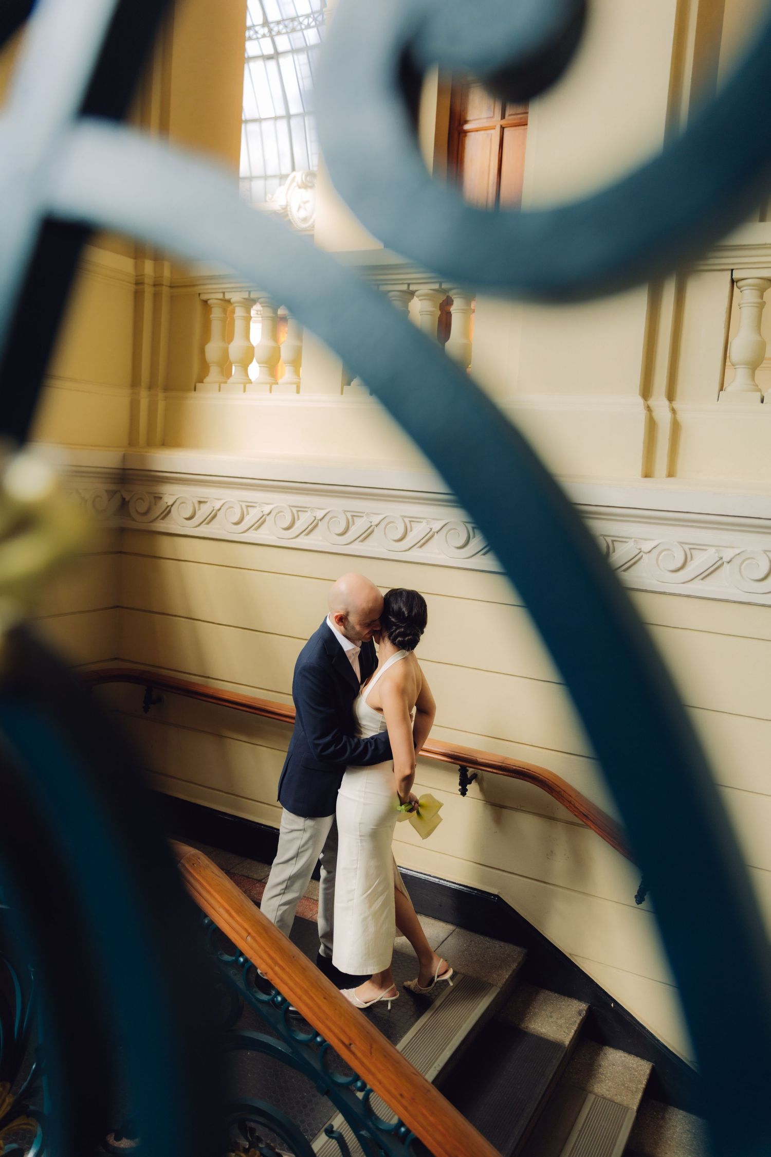 Bride and groom embrace on a grand staircase, captured through decorative blue wrought iron scrollwork railings.