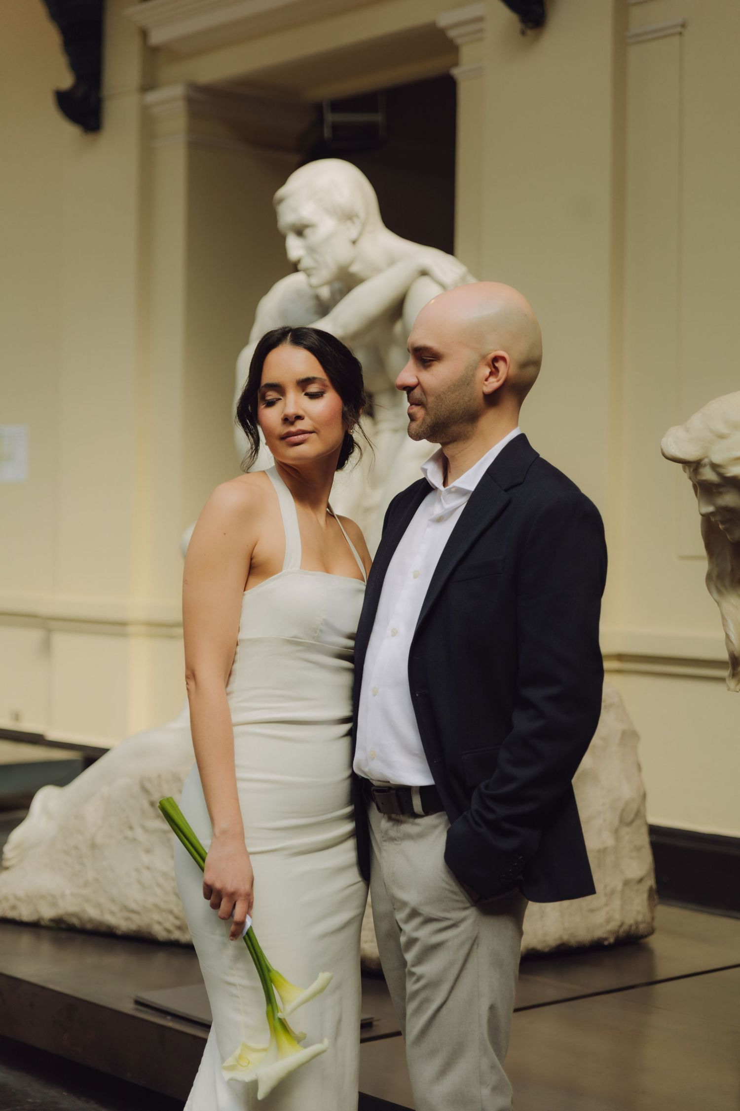 Bride in white halter gown and groom in navy blazer pose before classical marble sculptures in an art museum.