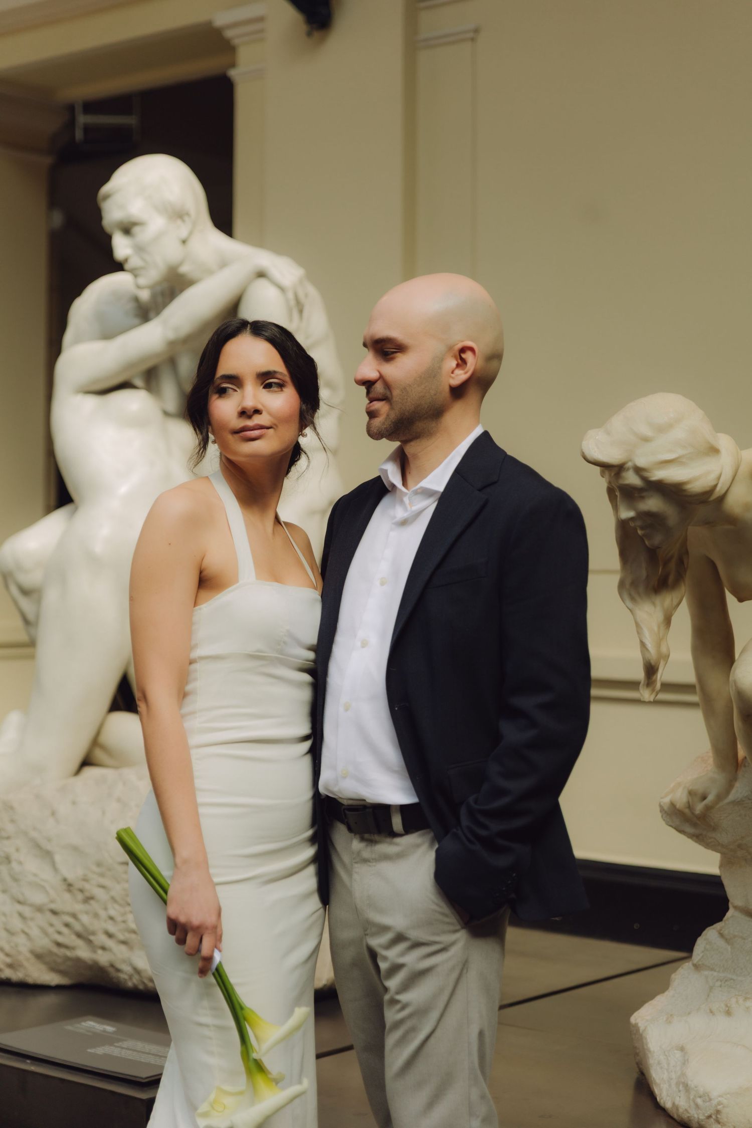 Elegant bride and groom stand together surrounded by classical white marble sculptures inside a grand art museum.