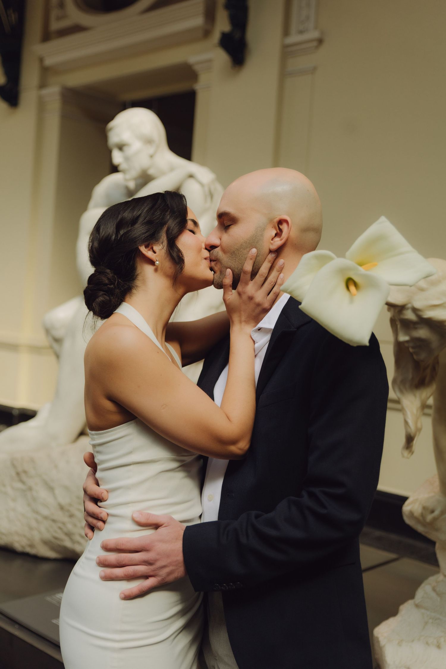 Newlyweds share a tender kiss surrounded by white marble statues in an elegant museum setting.