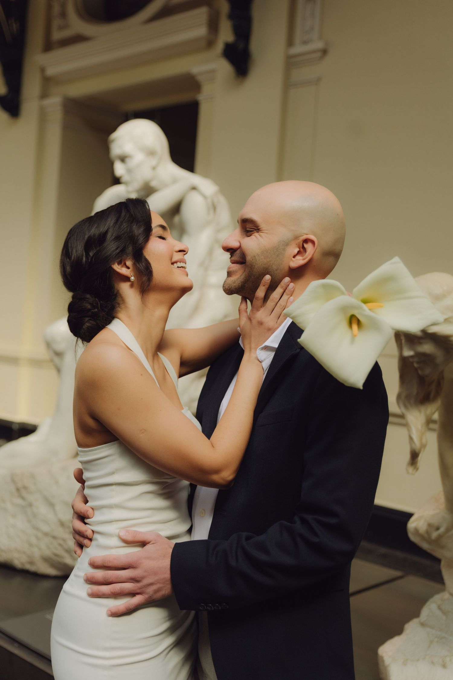 Smiling bride and groom embrace affectionately amid classical sculptures in a warmly lit museum.