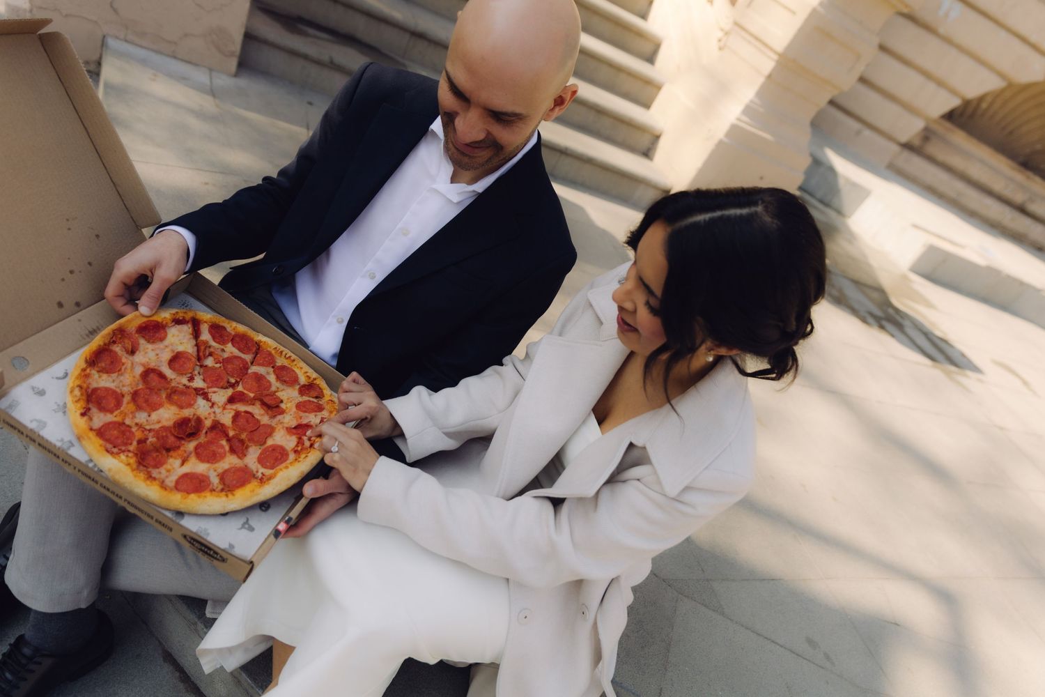 Bride and groom in wedding attire share a pepperoni pizza on outdoor steps of a grand building.