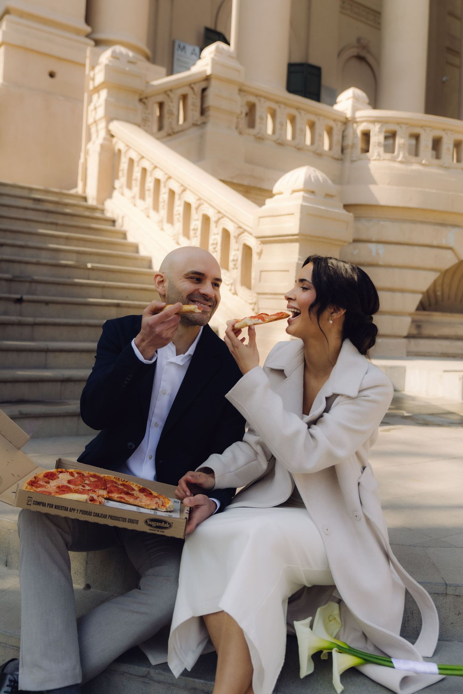 Elegantly dressed newlyweds laugh and eat pizza together on the steps of a neoclassical building.